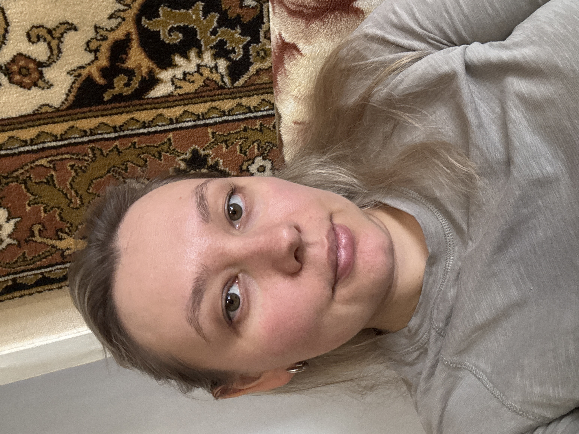 A close-up, chest-up image depicts an adult female in an indoor setting in Chaikovsky, Russia. The subject has light-toned skin, dark eyes, and light brown or blonde hair, partially visible around her shoulders. She wears a light grey, long-sleeved marled knit shirt with visible stitching at the neckline and shoulder. Small, silver hoop earrings are present on her left ear. Her lips are slightly parted, and she maintains direct eye contact with the viewer. The background features a patterned rug or tapestry with brown, gold, black, and cream motifs positioned to her right, and a plain light-colored wall or surface to her left. A light-colored, patterned textile, possibly a cushion or blanket, is partially visible to the right of her shoulder.