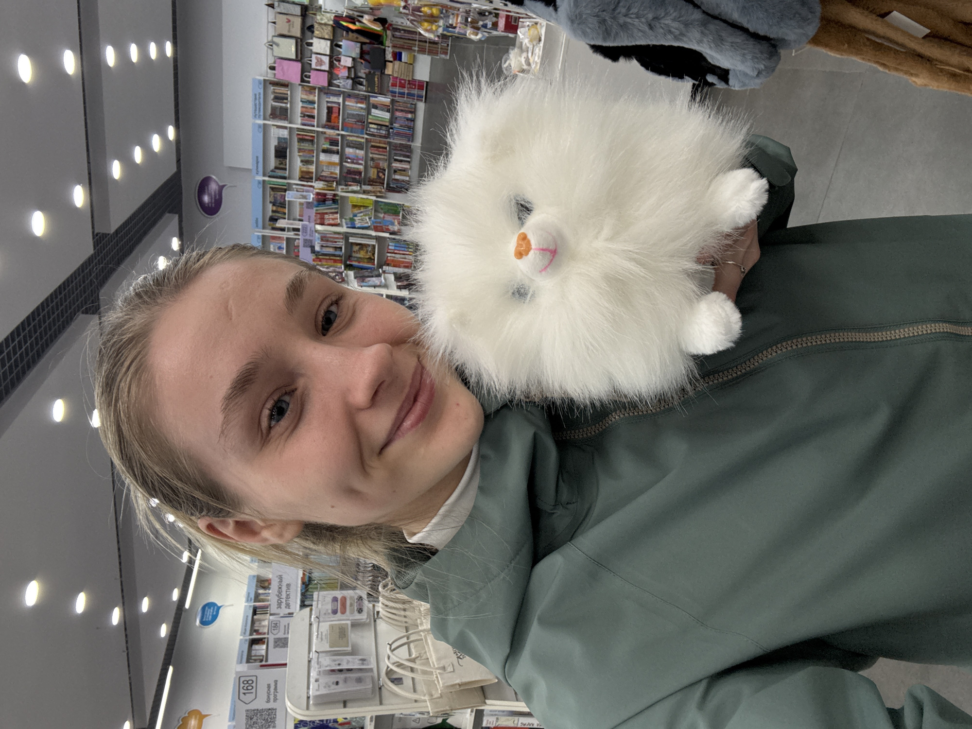 A person with fair skin and blonde hair is holding a fluffy, white, round animal plush toy. The plush toy has small paws, a pink mouth, a black nose, and black eyes. The person is wearing a green jacket with a white turtleneck underneath. The background shows shelves stocked with books, merchandise, and lights on the ceiling. Based on information from the provided context, this scene is likely located in a store in Izhevsk, Russia.