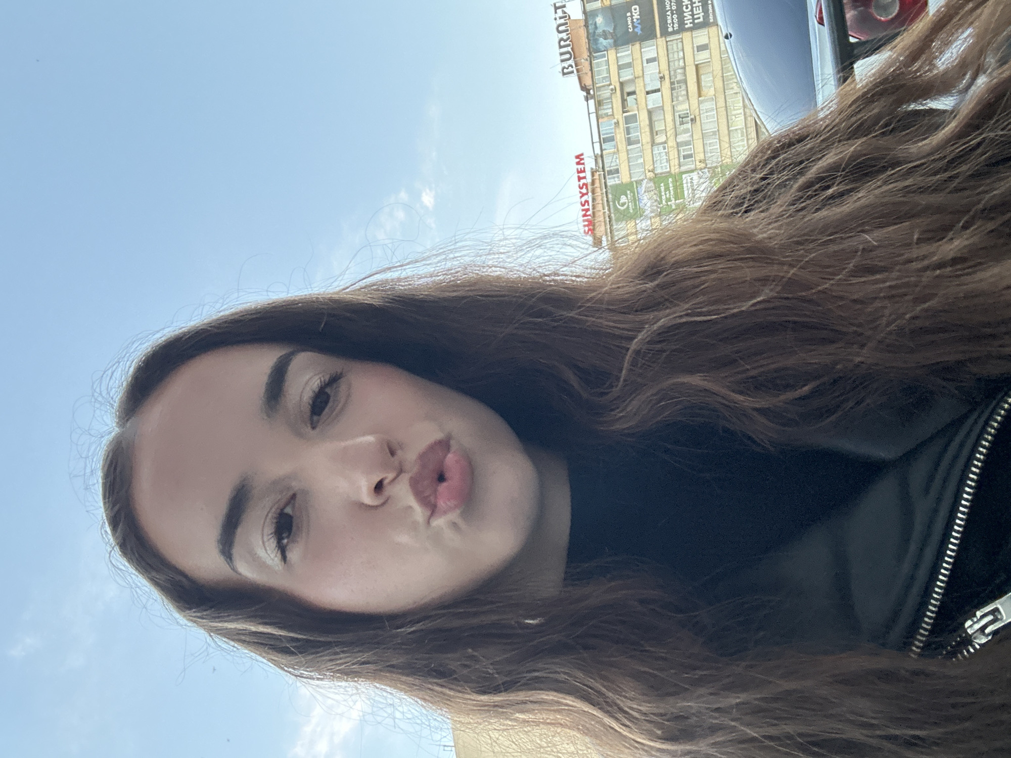 The image shows a young woman with long brown hair, pouting her lips, taking a selfie outdoors. She is wearing a black top and a black jacket with a silver zipper. In the background, there is a blue sky with some clouds, and a building with multiple windows. The building has several signs on it, including "SUNSYSTEM" and "BURDIT". There are also advertisements on the building, one of which appears to be for a car. The scene appears to be located in the city of Shumen, Bulgaria, as indicated by the text on the advertisements. The overall lighting suggests it is daytime.
