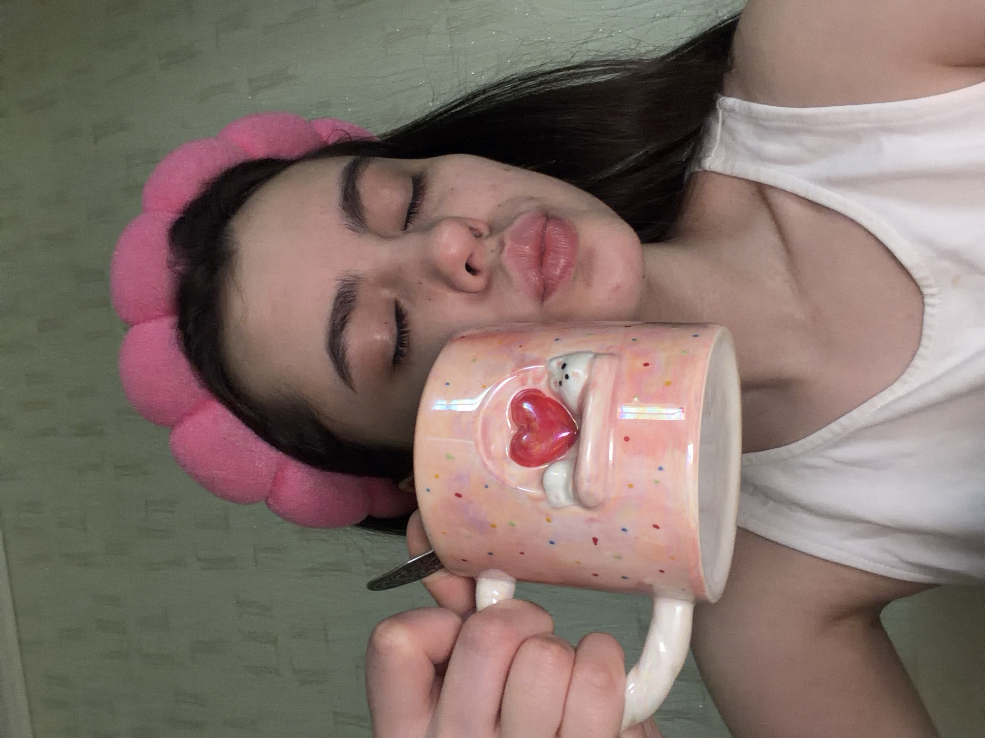 A young woman lies on her back, holding a mug, likely in a residential interior. The mug is light pink with colorful confetti dots and features a white bear design holding a red heart. She has dark hair and a pink headband. She is wearing a white tank top. The background suggests an interior with light green walls. There's no information in the image to confirm a specific location, such as Chaikovsky, Russia.