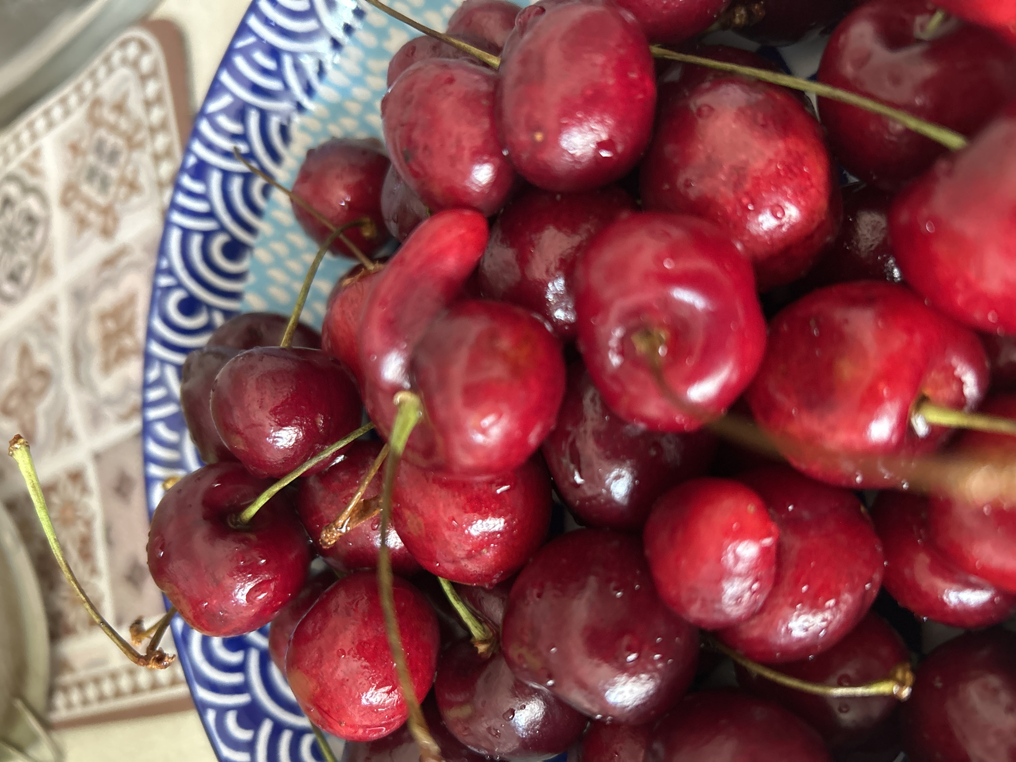 A close-up, overhead shot of a blue and white patterned bowl filled with fresh, dark red cherries. The cherries are plump and glistening with water droplets, and many still have their green stems attached. The bowl itself has a decorative rim with a blue wave-like pattern. In the background, a patterned placemat is visible. The overall impression is one of freshness and abundance. There is no visible indication of the city of Angarsk, Russia.