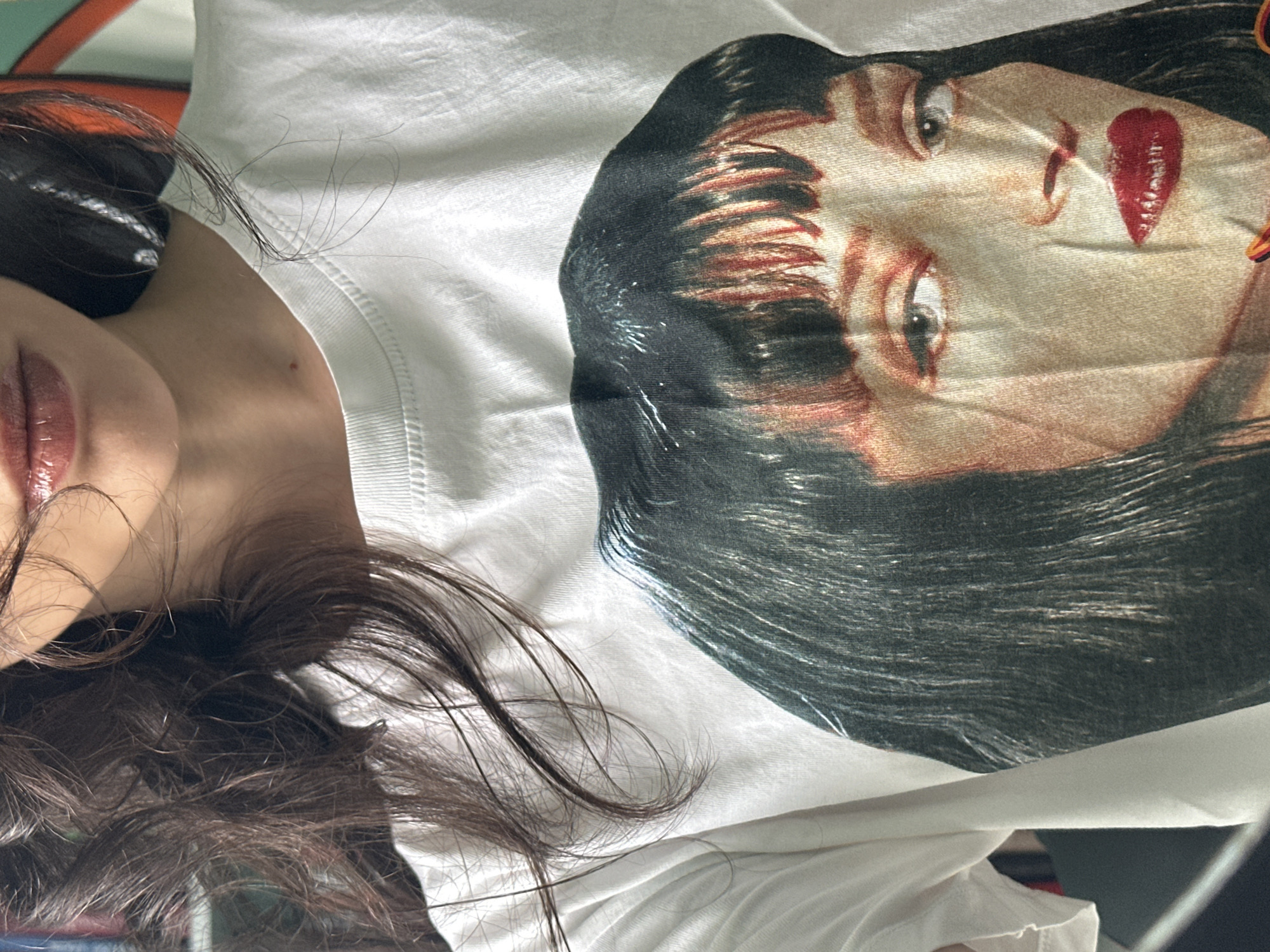 The media file shows a close-up, top-down view of a person wearing a white t-shirt with a graphic print of a woman's face. The person's dark hair is spread across the t-shirt and their left shoulder. The graphic on the t-shirt is a stylized depiction of a woman with dark hair, prominent eyes, and red lipstick, with a large red heart shape on her cheek. The lighting suggests an indoor setting, possibly a bedroom. No specific time of day or weather conditions are discernible. The image's composition focuses on the t-shirt graphic and the person's hair and upper body. The overall mood is neutral. No visible text is present in the image.