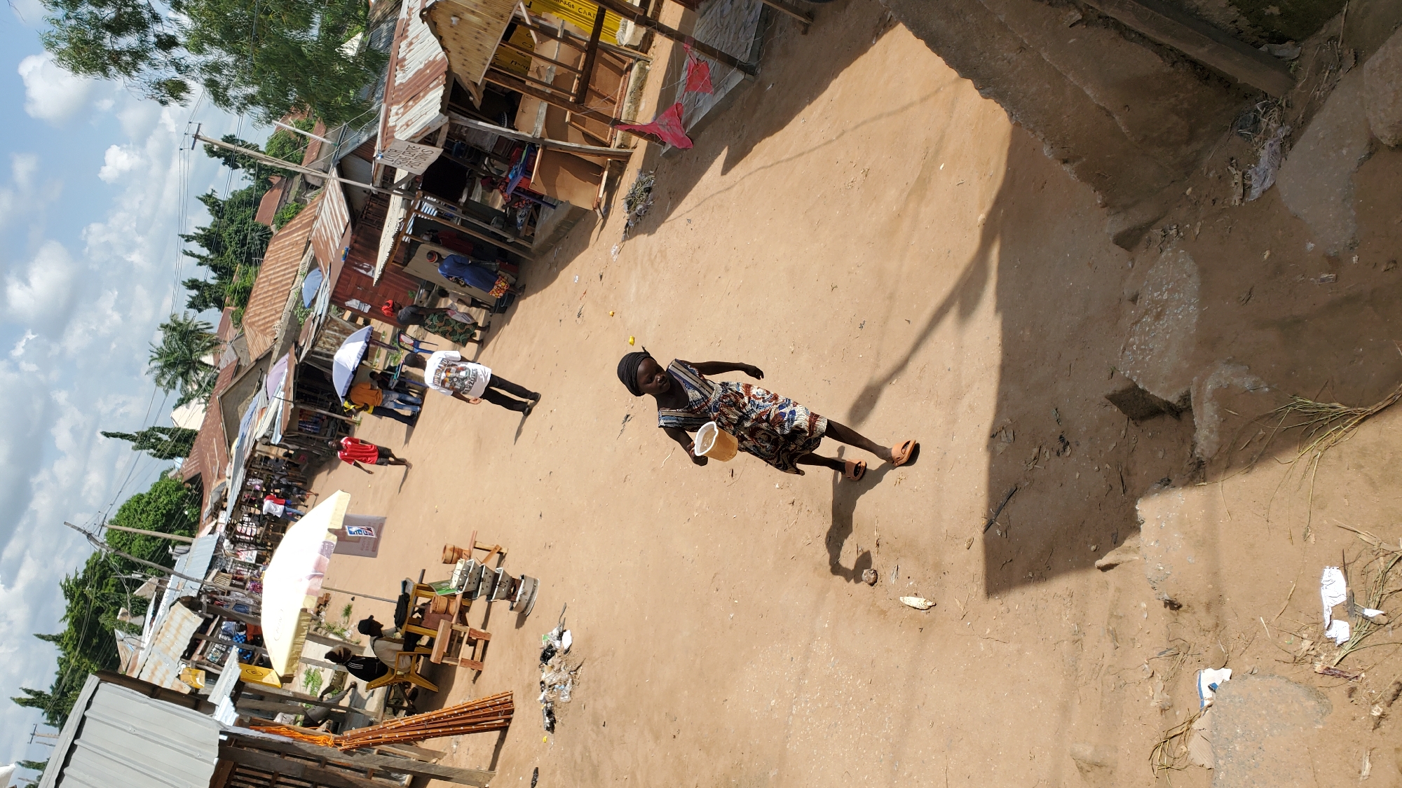 The image depicts a street scene in Jalingo, Nigeria. A young girl wearing a patterned dress and a black headscarf walks towards the viewer, carrying a light-colored bucket. She is wearing orange sandals. Behind her, in the middle ground, several people are visible, some standing under umbrellas and awnings. There are market stalls with corrugated metal roofs, suggesting a commercial area. In the background, trees and buildings line the street. The ground appears to be unpaved dirt. The lighting indicates it is daytime, with distinct shadows cast by the people and structures.