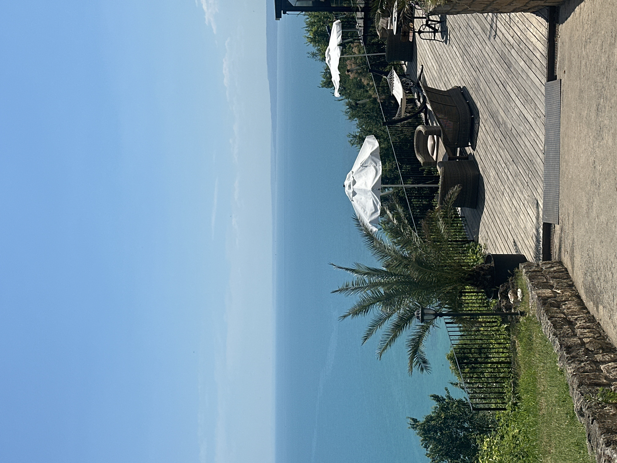 The image shows an outdoor seating area overlooking a vast blue body of water under a clear sky. The foreground features a rustic stone wall and a paved pathway leading down. To the left, lush green grass and foliage are visible, bordered by a black metal fence. Beyond the fence, a palm tree stands in a dark pot, next to a lamppost.
The main seating area is on a wooden deck. There are several chairs and tables, some with umbrellas providing shade. Specifically, there are two large white umbrellas, one closer to the foreground and another further back. Wicker chairs and sofas are arranged around tables. A hammock is also visible on the deck. In the background, dense green trees line the slope leading down to the water, and a portion of a building with dark-tinted windows can be seen on the far right. The water stretches to the horizon, with a faint line of land visible in the far distance. The scene appears to be a tranquil, scenic spot, likely a restaurant or cafe with a view. There are no people present in the image.
