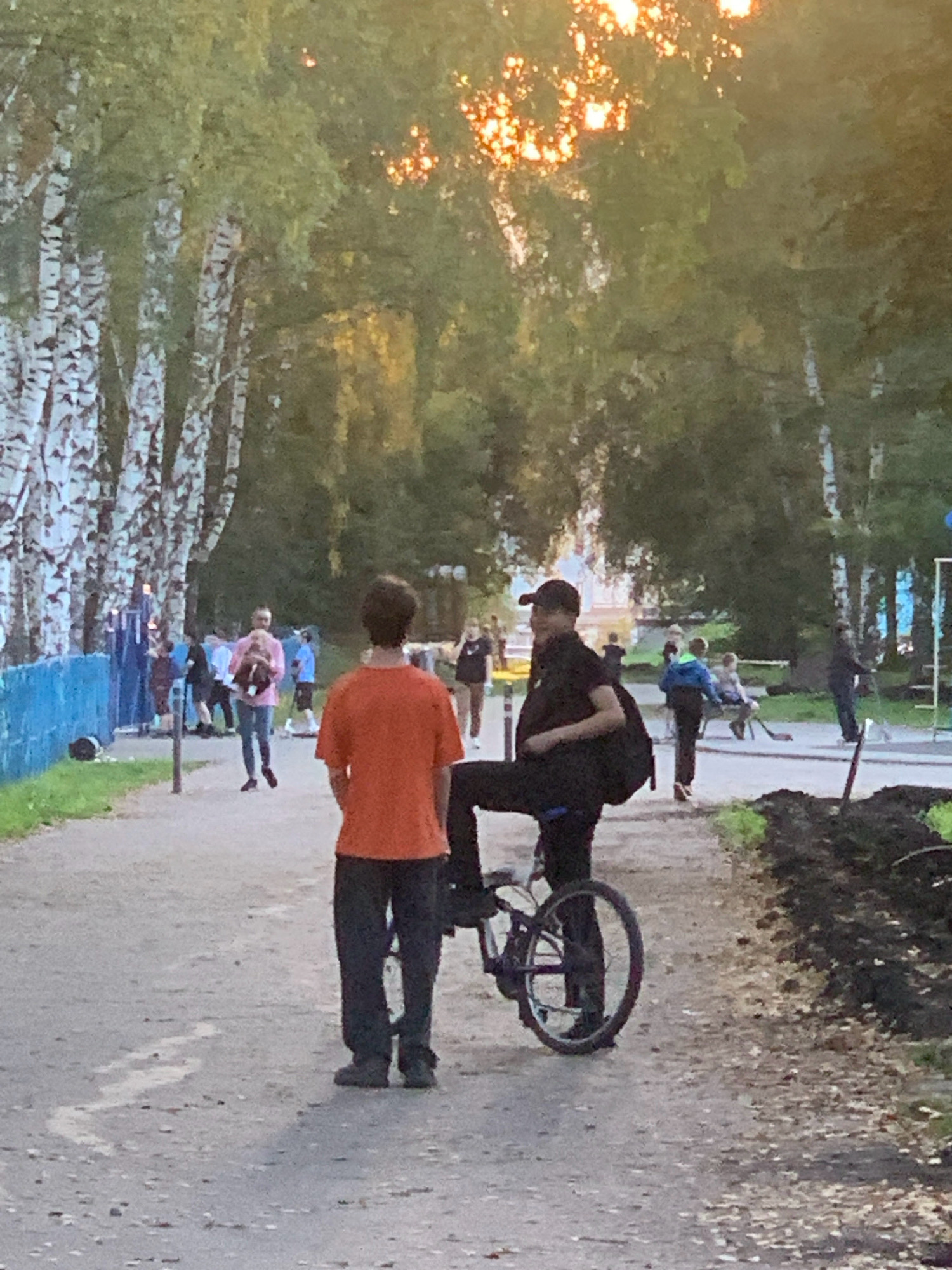 In Prokopyevsk, Russia, two young men stand on a dirt path lined with birch trees. The man on the left, wearing an orange t-shirt and dark pants, faces away from the camera, with his back to a group of people in the background near a blue fence. To his right, the second man, in a black t-shirt, black pants, and a black baseball cap, is seated on a bicycle, facing the man in orange. His left leg is extended, resting on the ground beside the bicycle's front wheel, while his right leg is bent, positioned over the bicycle frame. The bicycle has a black frame and black tires, and its rear wheel is clearly visible. Sunlight filters through the trees, casting dappled light on the path and the surrounding foliage, suggesting it is late afternoon or early evening. In the background, other individuals are scattered, some walking, others standing, and a few appear to be children playing or observing. A blue fence runs along the left side of the path, separating it from an unseen area. The ground is a mix of dirt and gravel, with some patches of grass and leaves visible, especially on the right side of the path where there is also disturbed earth, possibly indicating recent landscaping work.