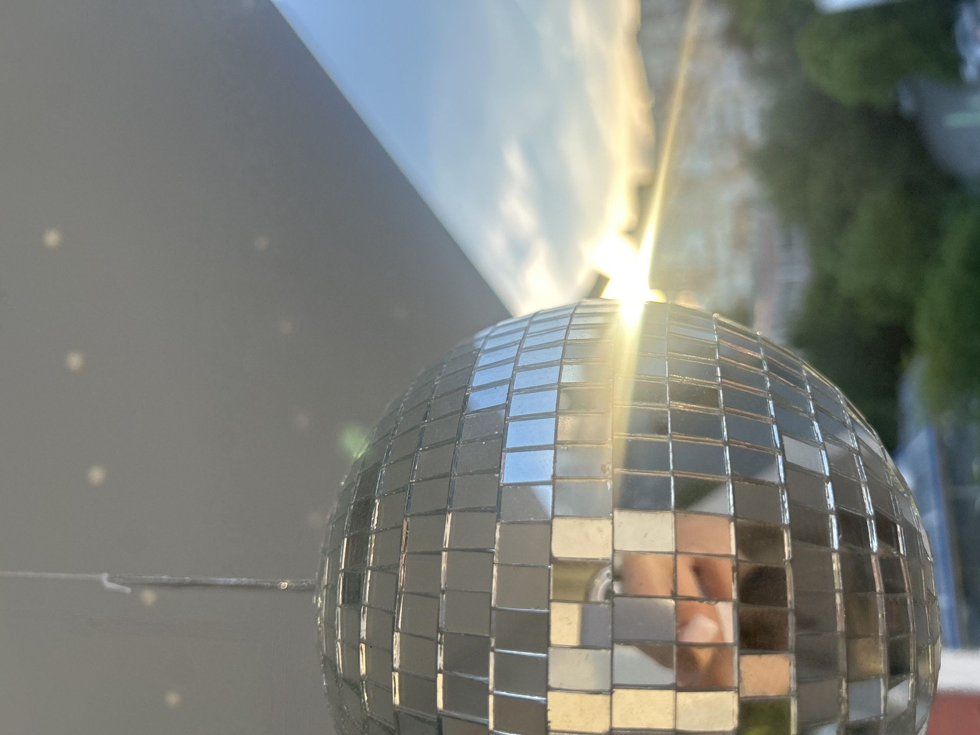 The image features a disco ball in close-up, reflecting a scene that includes the sky with sunlight and a building with greenery. The disco ball is made of small, mirrored squares. The reflection in the disco ball reveals a building, possibly residential, with green foliage suggestive of trees or shrubs. The sky is visible at the top of the image, with bright sunlight creating a flare effect. The disco ball is on a surface with light and reflections. Based on the visual evidence of urban architecture, natural light and reflection, and the presence of a disco ball, the location could be Veliko Tarnovo, Bulgaria.