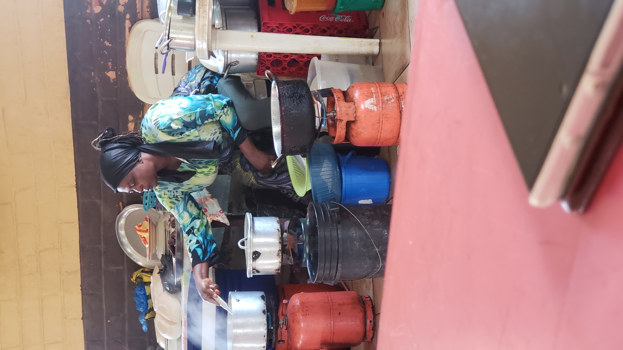 In Unguwar Shanu, Nigeria, a woman wearing a black headscarf and a blue and green patterned tunic is cooking outdoors. She is seated on a low stool, stirring a pot with a utensil. Several large metal pots and gas cylinders are arranged around her. Two red gas cylinders and one orange gas cylinder are visible, with pots placed on top of the cylinders to be heated. There are also several plastic buckets and basins nearby, some containing water or food items. In the background, a brick wall and wooden beams form the structure of the outdoor cooking area. The foreground is dominated by a red surface, possibly a table or a mat, with a smartphone resting on it. The scene suggests a communal cooking area or a busy street food vendor.