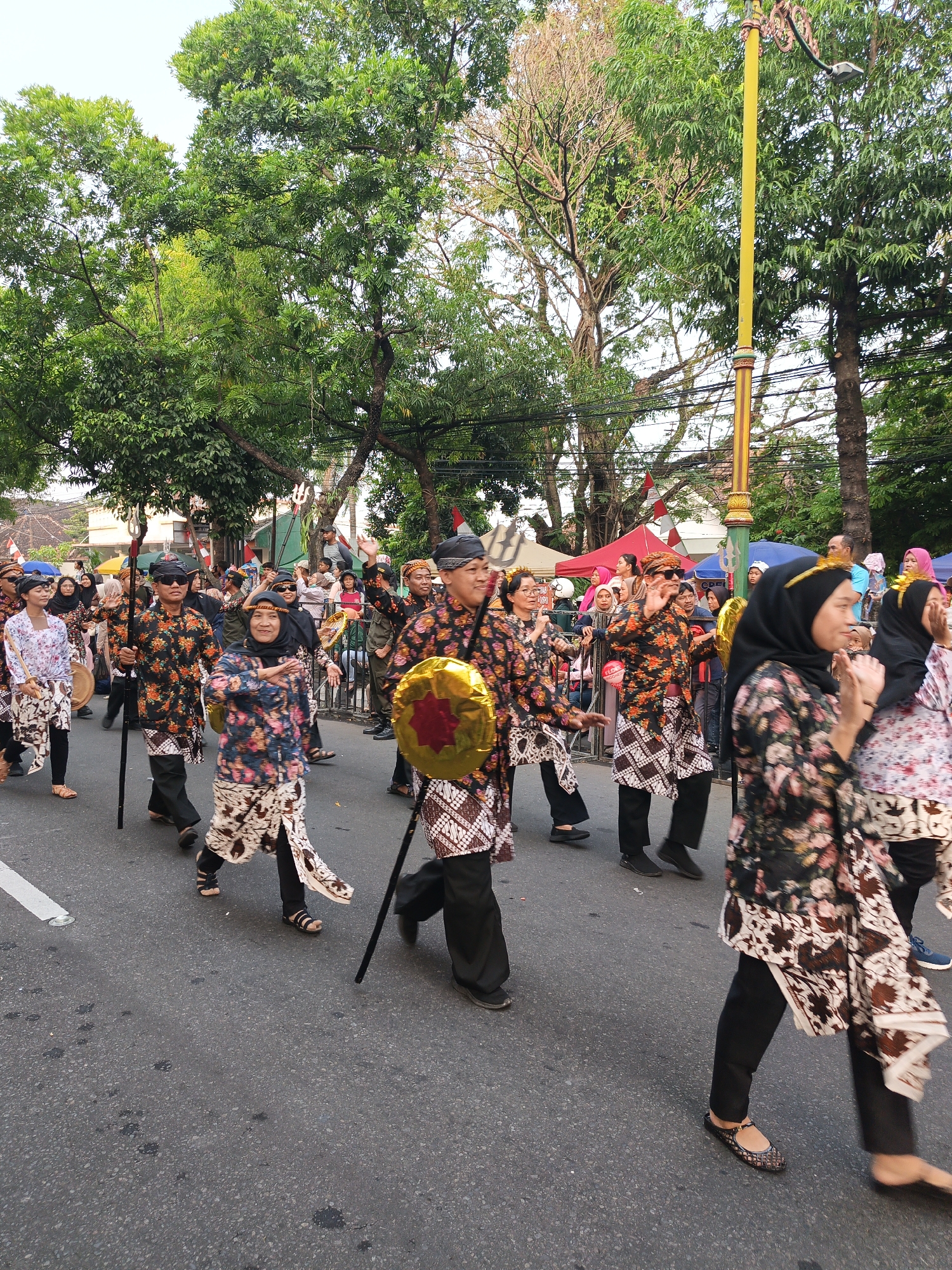 A group of people in traditional attire are participating in a parade on a street in Gayamprit, Indonesia. The individuals are wearing patterned shirts and skirts, some with headscarves. They are walking in the same direction, with some holding staffs or shields. Spectators are visible in the background, and trees line the street. The overall scene suggests a cultural event or celebration.