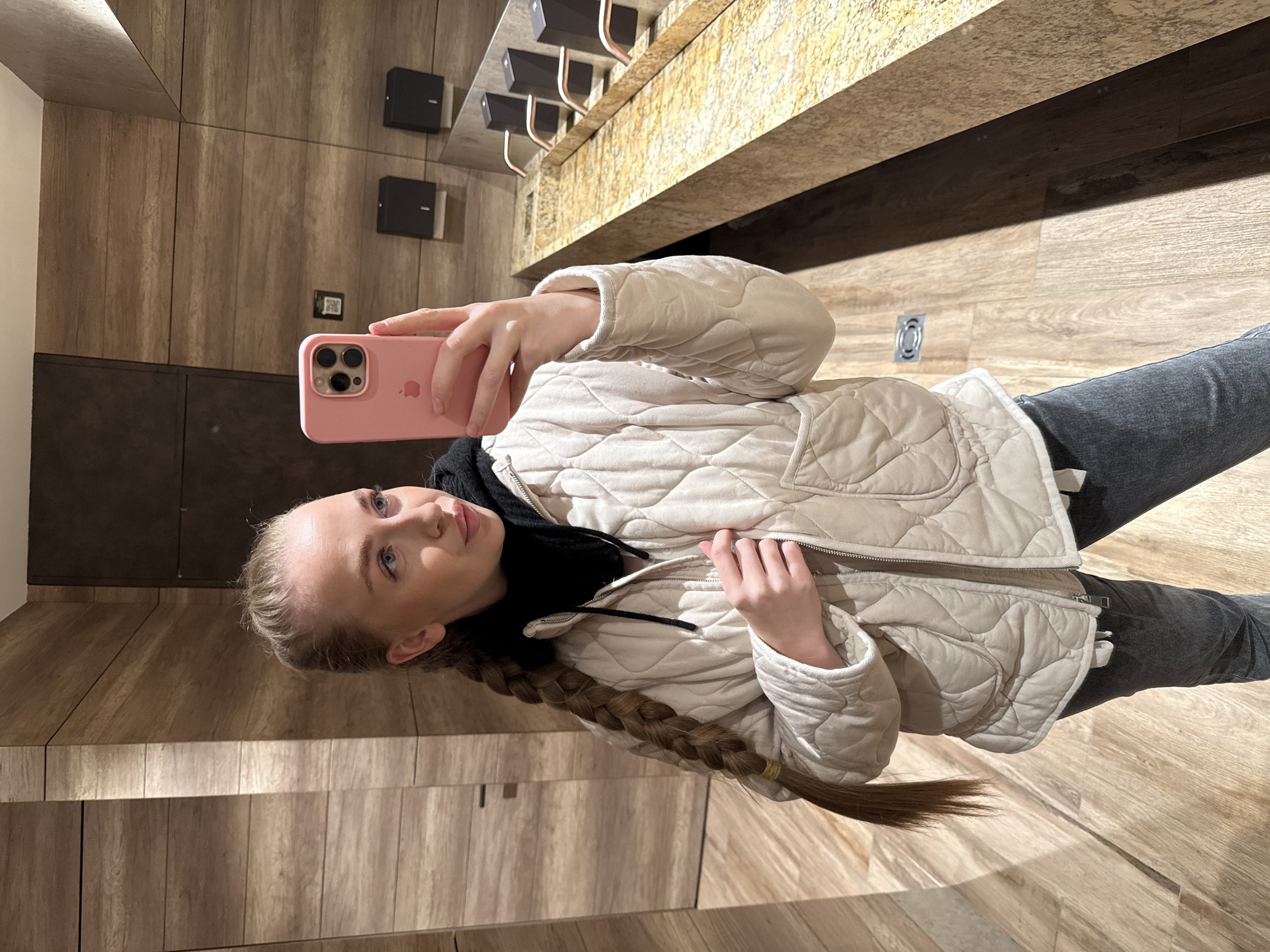 A young woman is taking a selfie in what appears to be a restroom or vanity area. She is holding a pink-cased iPhone in her right hand, angling the camera towards herself. She is wearing a beige quilted jacket over a black hooded top, and grey jeans. Her blonde hair is styled in a long braid. The backdrop features a wooden wall and a countertop with a modern design, including dark rectangular fixtures and copper-colored pipes. The lighting is ambient and warm. The location is Unknown, Unknown.