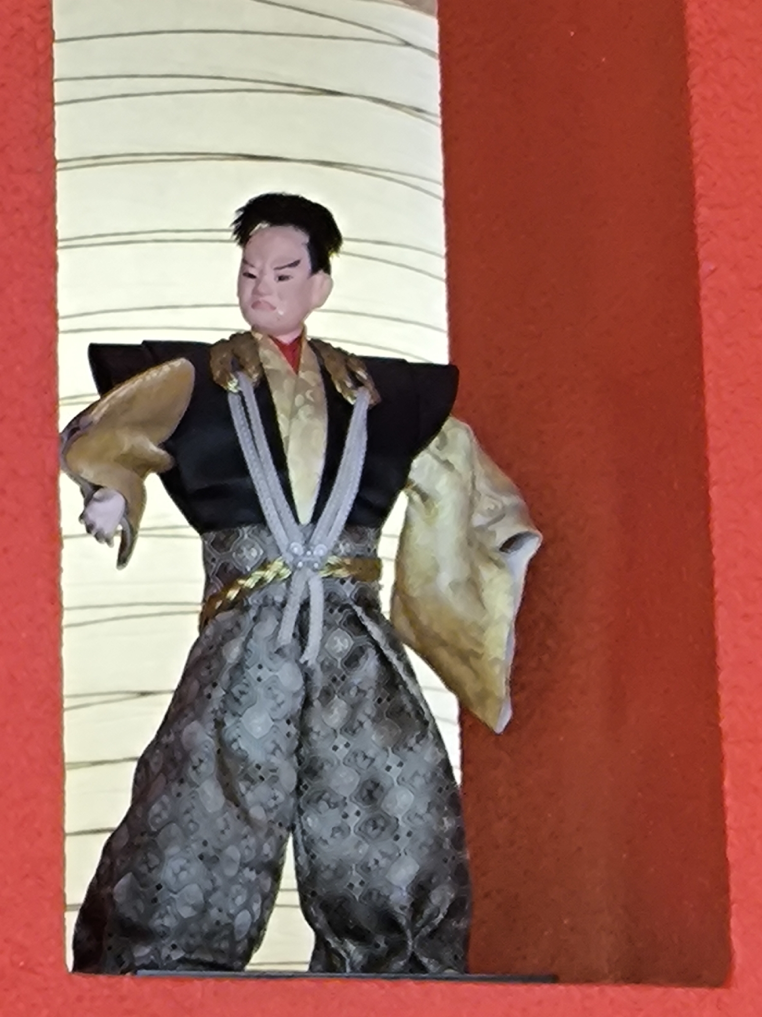 The image shows a traditional Japanese Kabuki doll displayed in what appears to be a niche or alcove, likely in an indoor setting. The doll is dressed in elaborate attire reminiscent of a samurai or noble character, featuring a black top with wide shoulders, an ornate gold and gray belt, and patterned hakama pants. The doll has an expressive face with dark hair styled in a traditional manner. The alcove has a red frame, and a white and yellow background suggests a lantern or screen. The image lacks specific information to confirm the location is Moscow, Russia, but the subject matter would indicate a museum or cultural display.