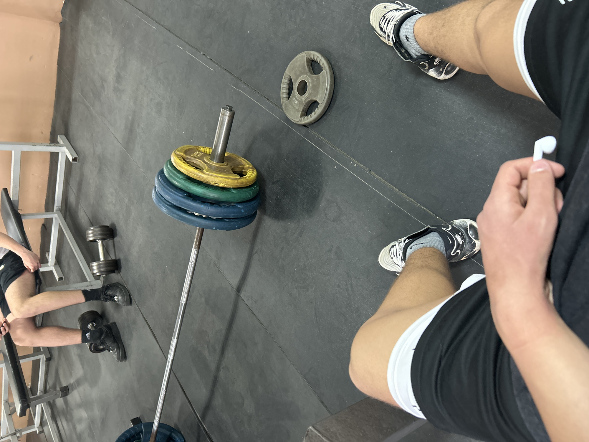 This image, captured from a high, tilted perspective, depicts an indoor gym setting. The floor is covered in dark gray rubber matting, and a light beige or pale orange wall is visible in the background on the left.
Two individuals are partially visible. In the immediate foreground, a person's legs and right arm are shown. They are wearing black shorts with a white waistband, gray socks, and black and white Nike sneakers, clearly displaying the "NIKE" logo and swoosh. Their right hand holds a white wireless earbud, suggesting they are listening to music or are about to.
To the left, another person's legs and lower torso are visible, positioned as if sitting on a piece of gym equipment, possibly a bench. This individual wears black shorts, black socks, and black sneakers, one of which shows a visible brand logo that appears to be Adidas. Near this person, a set of dumbbells rests on the floor.
The central focus of the equipment is a barbell standing upright, loaded with several colored weight plates. From top to bottom, there is a yellow plate, followed by at least one green plate, and then multiple blue plates. A separate, single gray weight plate, marked "10KG", lies flat on the rubber floor further into the scene, near the foreground person's feet. The overall scene suggests a pause or rest period during a weightlifting workout session at a gym in Zlatograd, Bulgaria.
