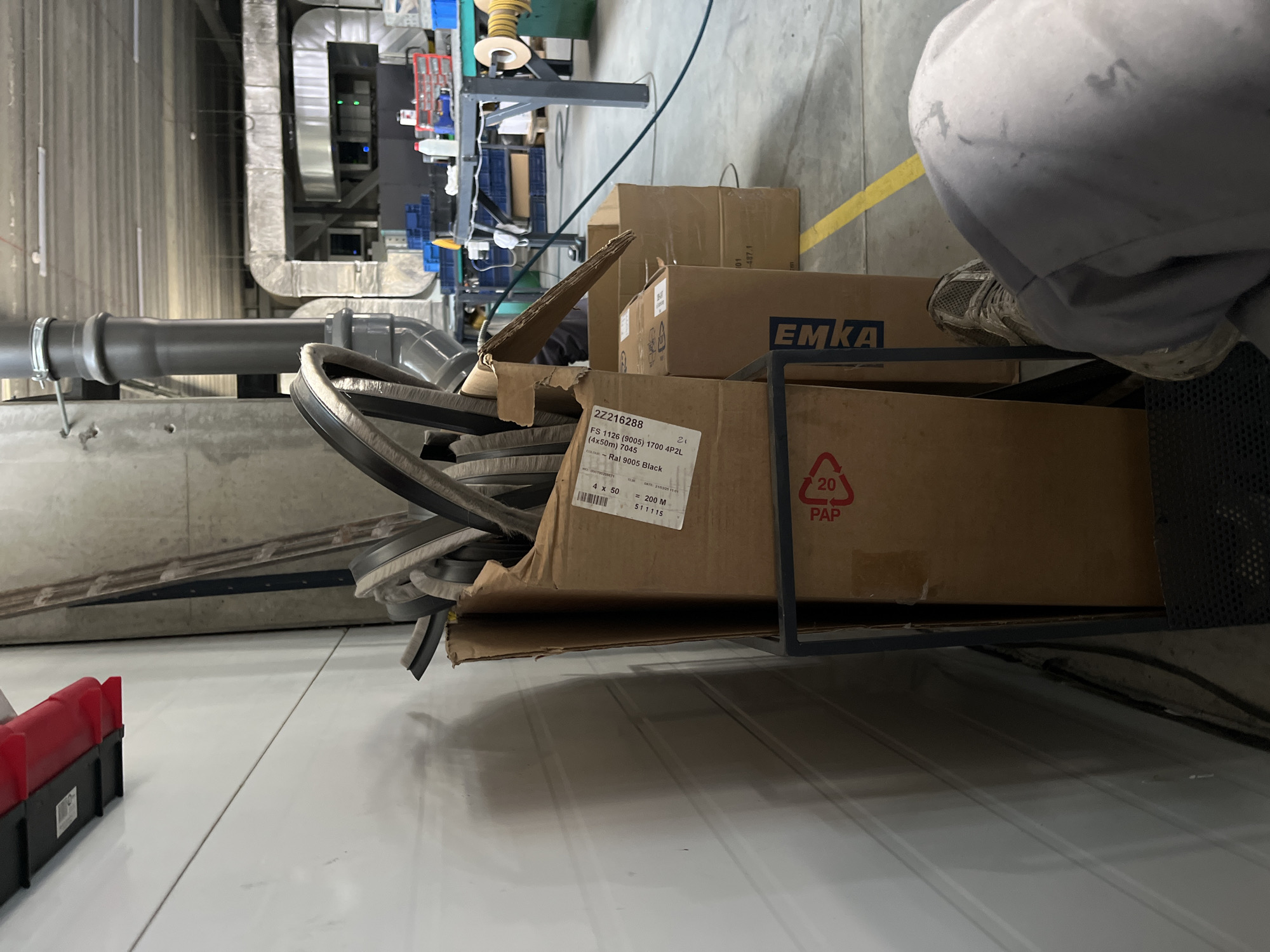 An interior shot of a warehouse in Kyiv, Ukraine, shows a tall, grey industrial pipe with a silver clamp, and ventilation ducts. In the foreground, a person's legs and feet are visible, one foot wearing a grey and black athletic shoe. The person is wearing light grey work pants that appear stained. To the left of the person, a tall, white corrugated metal wall is present. In the center of the image, several cardboard boxes are stacked, with the top box overflowing with what appear to be rubber or plastic seals with brush-like edges. One of the boxes has a label that reads "EMKA". To the right of the boxes, a yellow line is painted on the floor, and beyond that, a metal table with blue plastic bins, tools, and a spool of wire is visible. A green industrial machine is also present in the background.