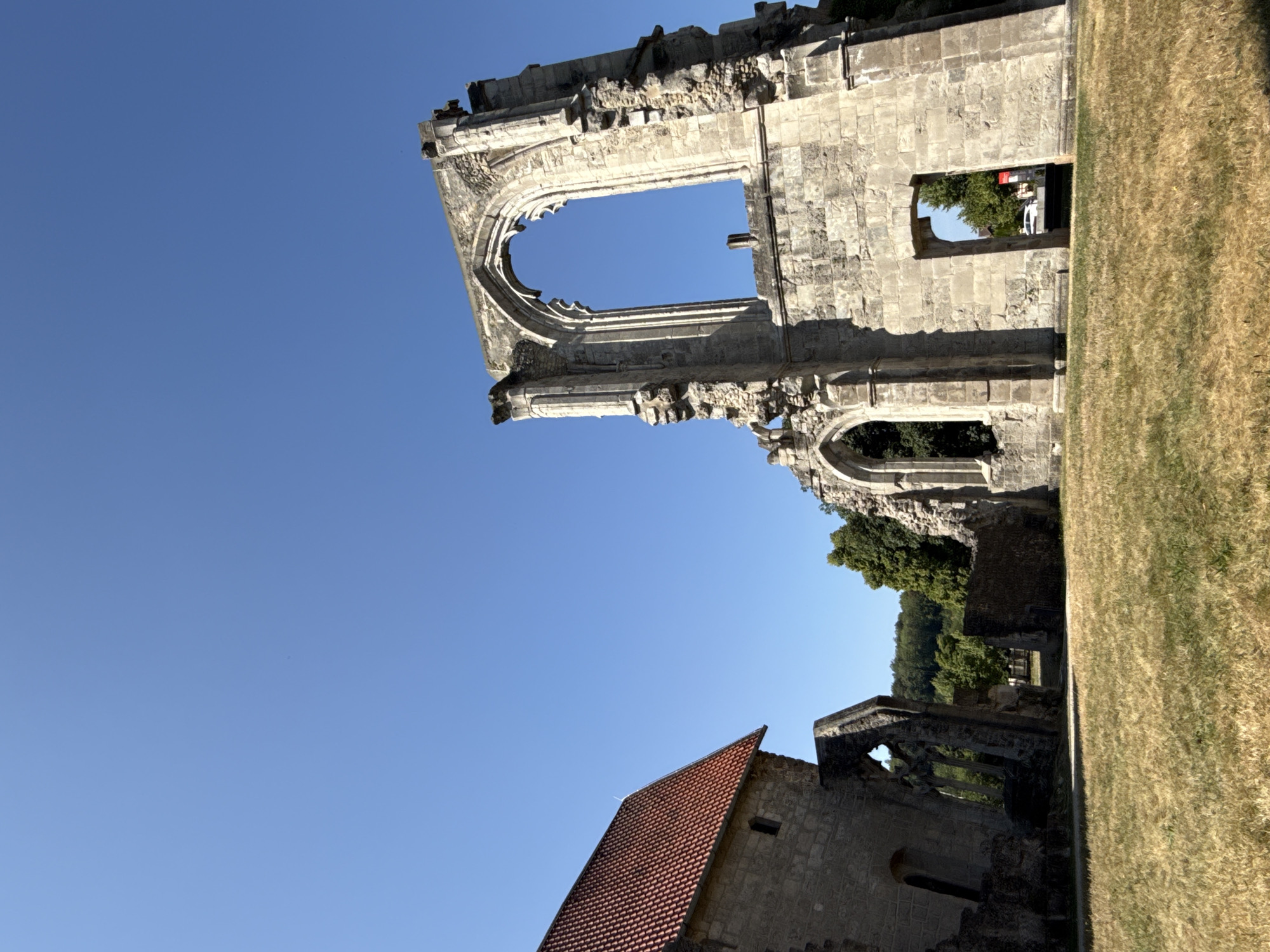The image shows the ruins of the Walkenried Abbey, located in Walkenried, Germany. The ruins are made of stone and are partially overgrown with vegetation. The sky is clear and blue. In the foreground, there is a grassy field. To the left, a stone building with a red tile roof stands intact. To the right, a large, arched window frame made of stone is the most prominent feature of the ruins. The abbey was founded in 1137 and was one of the most important Cistercian monasteries in Germany. It was dissolved in 1803 and later partly demolished. Today, the ruins are a popular tourist attraction.