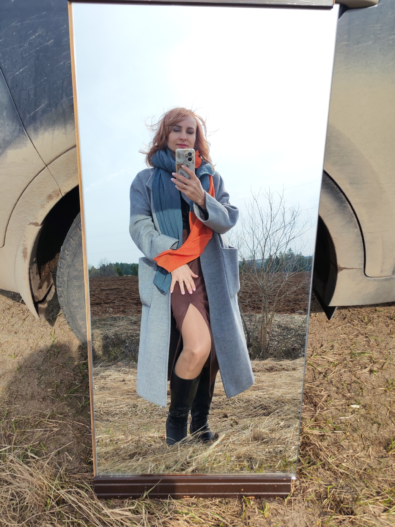 A fair-skinned woman with red hair stands in front of a full-length mirror in a rural environment, possibly near Нечкино, Russia, based on the landscape and vegetation. She is wearing a gray overcoat, a brown dress with a thigh-high slit, and black boots. A blue and orange scarf is draped around her neck. The woman is holding a smartphone in her right hand, presumably taking a selfie. The mirror's reflection shows a field with bare trees and a plowed area. The mirror is situated near the dirty wheel and fender of a dark-colored vehicle, suggesting the location is outdoors with the ground covered in dry grass and dirt. The sky is overcast with no visible sun.