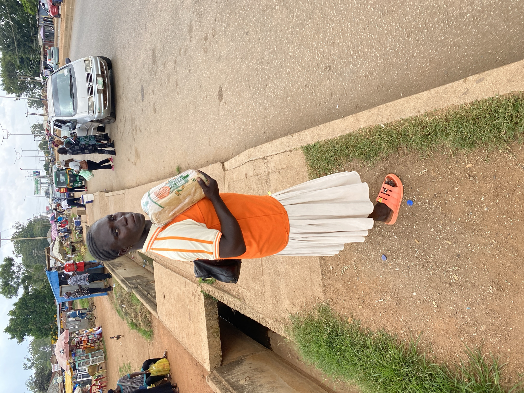 The image depicts a street scene in Bukuru, Nigeria, likely during the daytime given the ambient light. The foreground features a young woman wearing an orange and white striped shirt and a long white skirt. She is carrying a large loaf of bread wrapped in plastic over her shoulder. She is wearing bright orange sandals. She appears to be walking along a dirt and concrete path next to a drainage ditch. In the background, there are several stalls or shops set up along the roadside, with various goods on display, including food items and household products. A few vehicles, including a silver minivan and a tricycle (Keke Napep), are visible on the road. Other people are present, some appearing to be vendors and others possibly shoppers or passersby. The setting is an outdoor marketplace with a busy atmosphere. The ground is a mix of dirt and concrete, with some patches of grass. The overall impression is of a vibrant and active local commercial area.