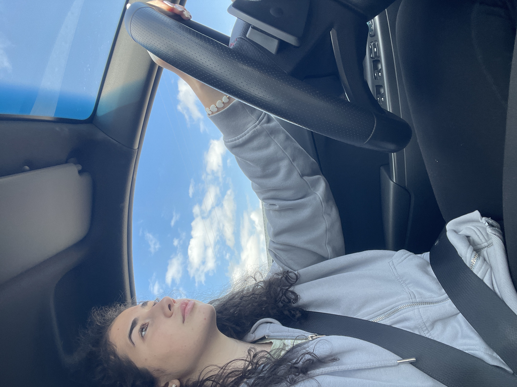 A young woman with curly dark hair, wearing a light gray hoodie and a seatbelt, reclines in the driver's seat of a car. Her face is tilted upward, gazing toward the sunroof, through which a bright blue sky with scattered white clouds is visible. Her left hand, adorned with a white beaded bracelet, rests on the steering wheel. To her right are the vehicle's window controls. The location cannot be determined based on this image.