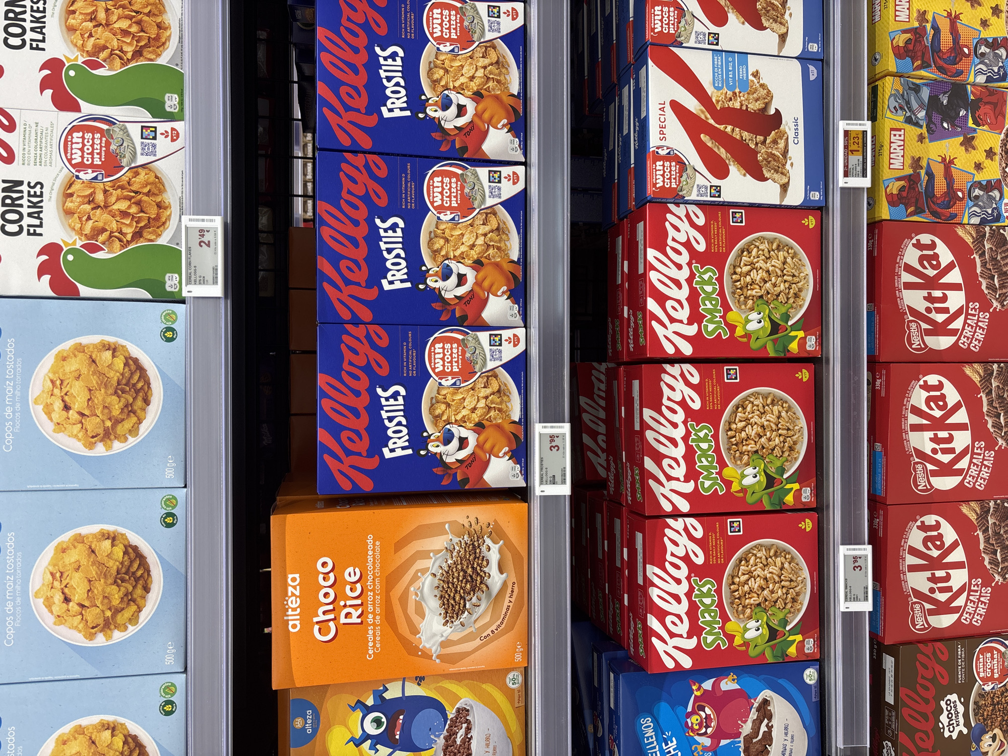 This is a mid-level shot of a supermarket aisle featuring various breakfast cereal boxes on shelves. The lighting is bright, indicating artificial store lighting. The image is focused on the products, with no people visible.
On the top shelf, from left to right, are several boxes of "Copos de maíz tostados" (toasted corn flakes). The packaging is predominantly light blue with an image of corn flakes in a white bowl. Some boxes have green circular logos.
The middle shelf showcases a variety of cereal boxes. On the far left is an orange box labeled "alteza Choco Rice Cereales de arroz chocolateado" with a cartoon monster illustration. To its right are several bright blue boxes of "Kellogg's Frosties" featuring Tony the Tiger. Below these, on the next shelf down, are red boxes of "Kellogg's Smacks" with a frog character. Next to the Smacks boxes are white and blue boxes of "Kellogg's Special K" with a red K logo.
The bottom shelf displays more cereal boxes. On the left are boxes of "Kellogg's Choco Krispies" with a monkey character. To their right are red and white boxes of "Nestlé Kit Kat Cereales Cereais" featuring images of chocolate. Further to the right are boxes of "Marvel" cereals with superhero graphics from the Marvel franchise.
A price tag for Kellogg's Corn Flakes is visible on the middle shelf, showing "2'49 €". Another price tag for Kellogg's Smacks reads "3'95 €". A promotional tag for "2ª UNIDAD -50%" (2nd unit -50%) is also visible on the bottom shelf with a price of "1,23 €". The arrangement of products suggests a well-stocked retail environment. The specific location within Urbanización Villa Martín, Spain, is not directly identifiable from the visual cues alone, but the presence of Spanish language on the packaging confirms a Spanish-speaking market.