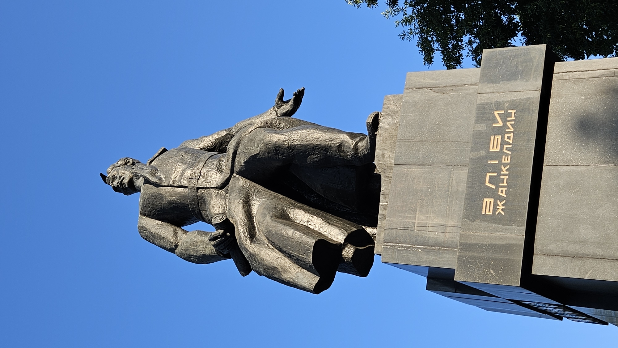 A large, dark grey granite monument featuring a statue of a man is situated in Almaty, Kazakhstan. The statue depicts a man in a long coat, seated with his left arm resting on his lap and his right arm extended outwards with an open palm. The man has a determined expression and appears to be looking upwards. The base of the monument has text inscribed in gold lettering that reads "ӘЛІБИ ЖАНКЕЛДИН". The monument is positioned outdoors against a clear blue sky, with a portion of a green tree visible on the right side of the frame. The image is taken from a low angle, looking up at the monument.