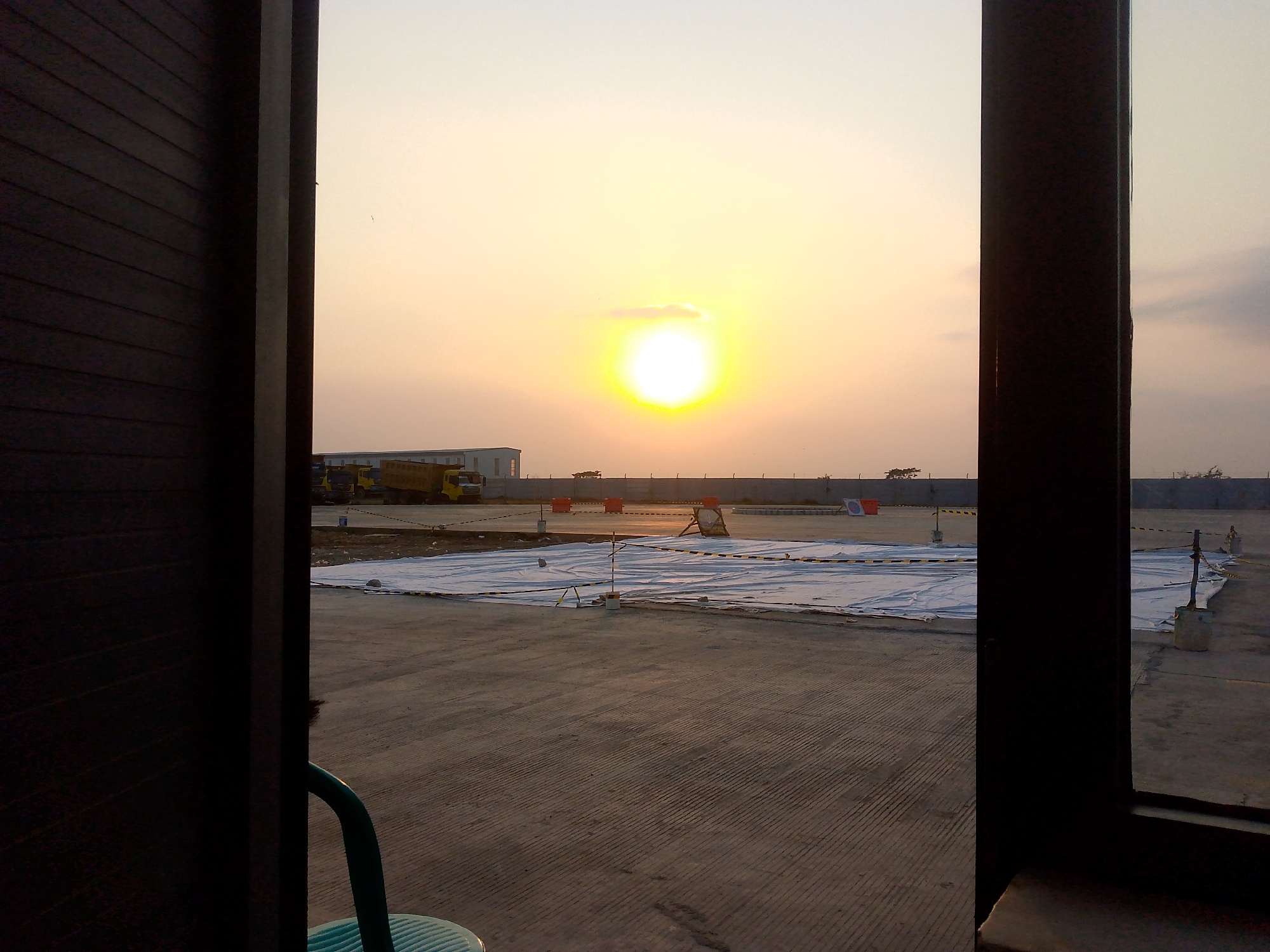The image captures a sunset scene from a construction site, likely in Krimun, Indonesia. The foreground is dominated by dark vertical structures framing a view of an open, paved area. In the middle ground, large white tarps are spread across the ground, secured with caution tape, suggesting ongoing construction or material protection. Yellow trucks are visible in the distance near a building. The sky is a bright orange and yellow gradient, characteristic of a sunset, with the sun prominently displayed as a bright, glowing orb. The environment appears industrial and unfinished.