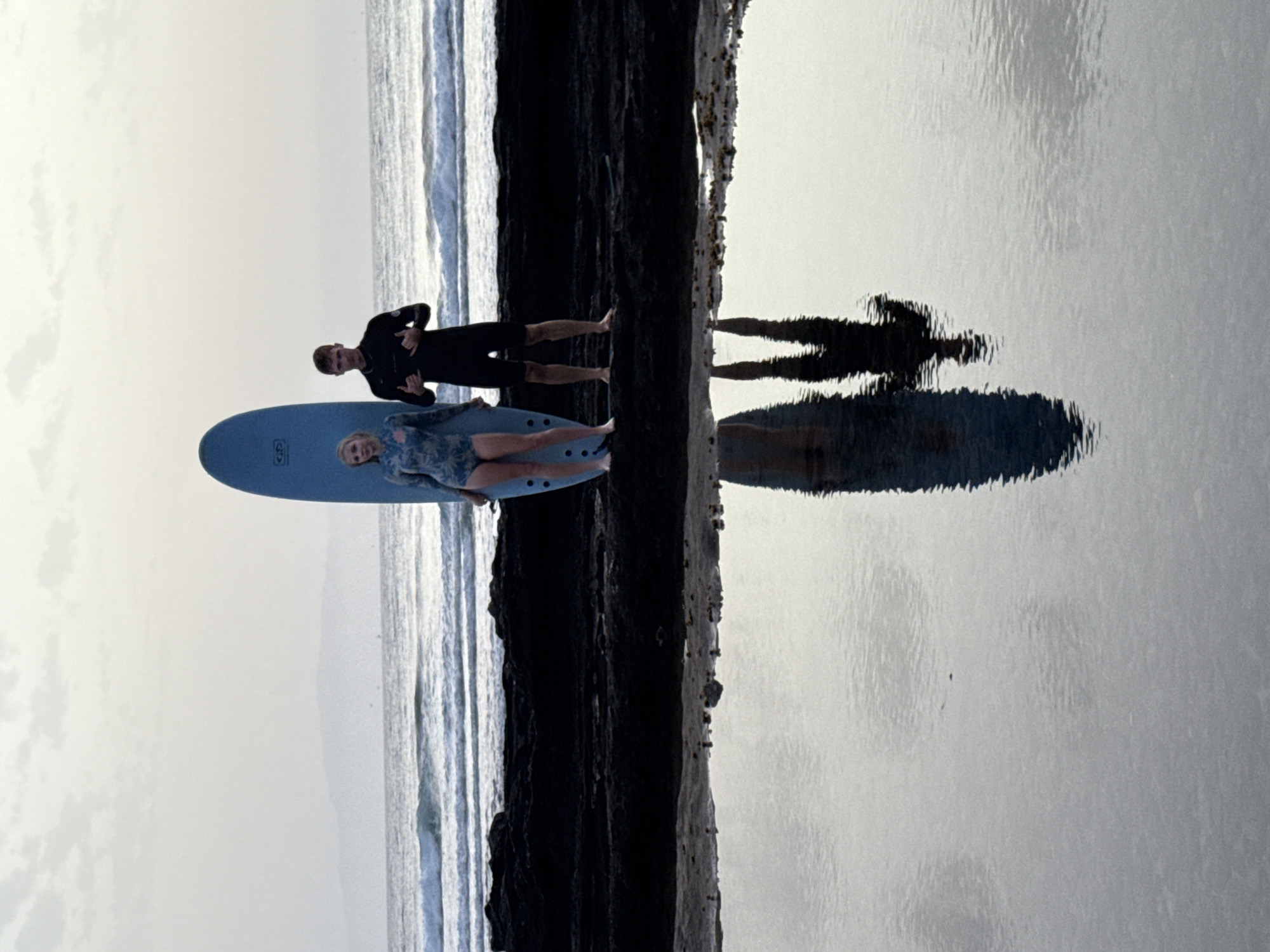 Two people, a woman and a man, stand on a dark, rocky shore with a surfboard standing upright between them. The woman, on the left, wears a long-sleeved swimsuit with a blue and white floral pattern. The man, on the right, wears a black wetsuit. They are both facing the camera. The ocean with small waves is visible behind them, and a distant, hazy coastline is in the background under a pale sky with scattered clouds. In the foreground, a calm pool of water reflects the figures and the surfboard. The scene appears to be on a beach near Arona, Spain, given the coastal landscape and the quality of light.