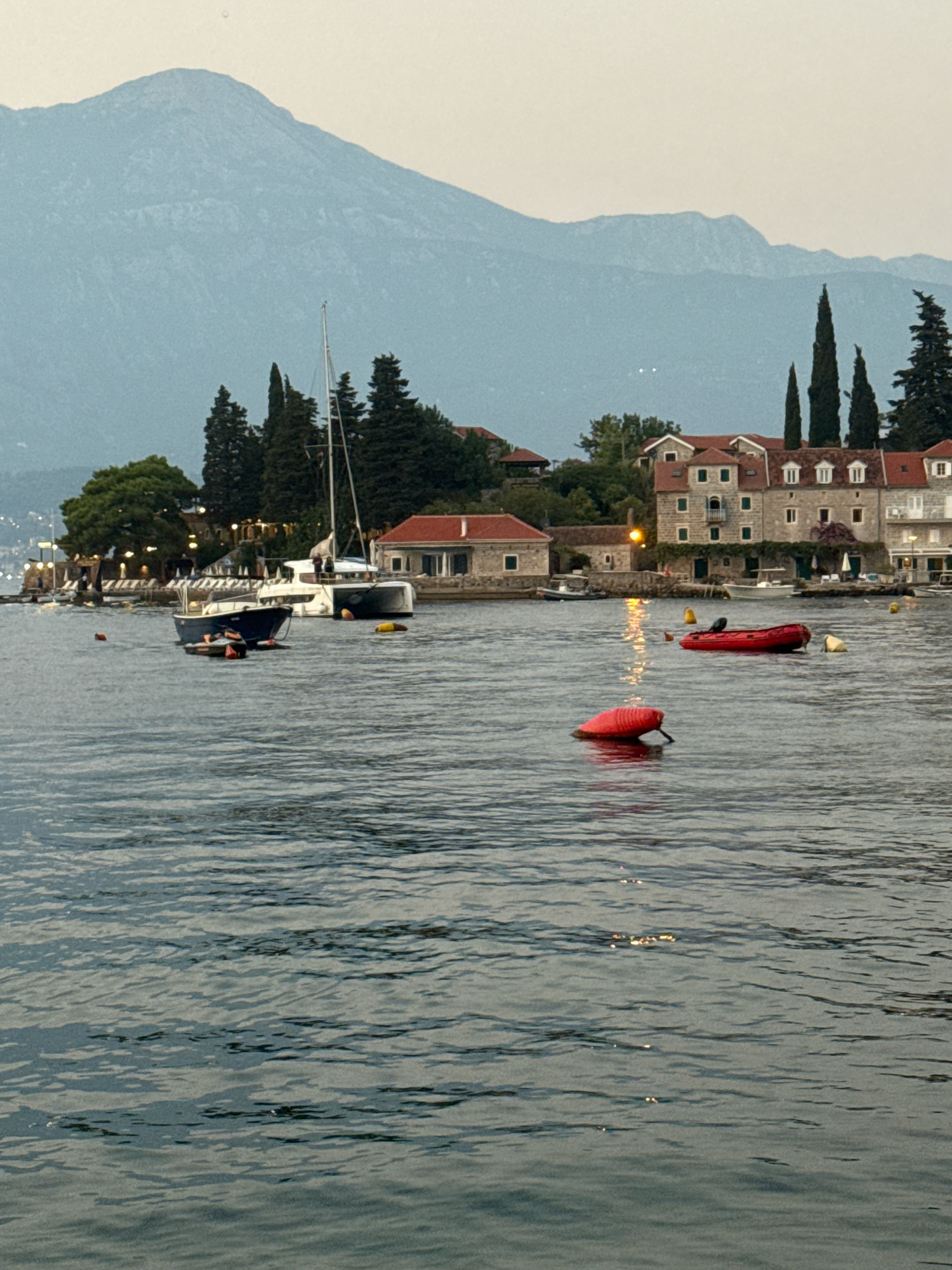 A scenic view of a bay with boats, buildings, and mountains in the background. In the foreground, the water is a deep blue with gentle ripples. Several buoys, one bright red and another orange, are visible on the water. A small blue boat with an orange fender is moored closer to the shore, and a larger white catamaran is anchored further out. Behind the boats, a cluster of buildings with red-tiled roofs and stone facades line the coast. Tall, dark green trees stand between the buildings and a body of water. In the distance, a mountain range with soft, hazy peaks rises against a pale sky, suggesting either early morning or late evening. The scene appears serene and tranquil.