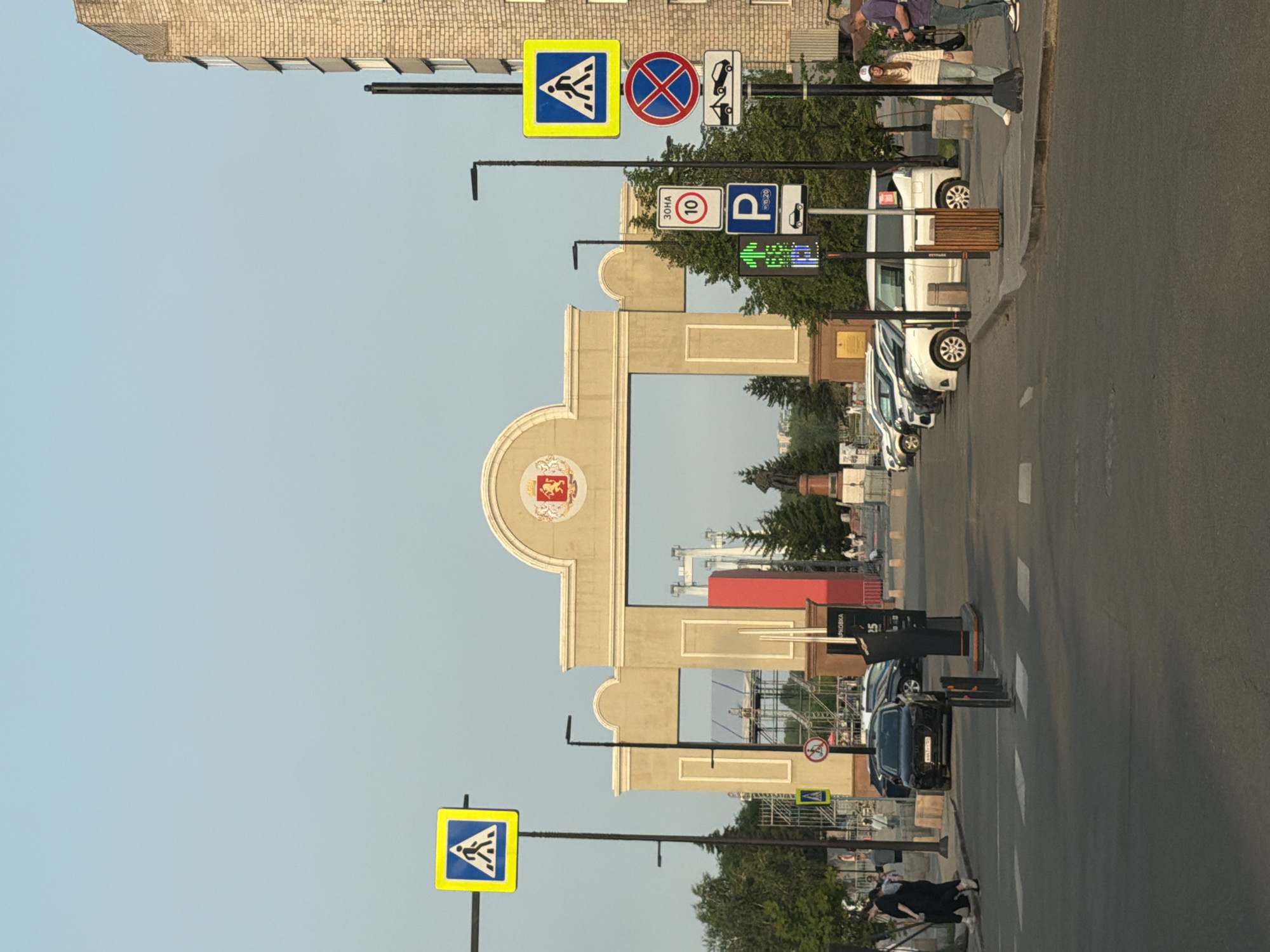 The image shows a wide, paved street in front of a large, ornate archway, possibly an entrance to a park or public space, located in Krasnoyarsk, Russia. The arch is made of light-colored stone with decorative moldings and a crest at its apex. Behind the arch, scaffolding and structures suggest ongoing construction or event setup. To the left and right of the arch, trees are visible, and on the far right, a tall, brick building stands.
Several traffic signs are prominently displayed on poles along the street. These include pedestrian crossing signs, a "ZONE 10" speed limit sign, a parking sign, a "no stopping" sign, and a towing sign. A traffic light with a pedestrian crossing signal is also visible.
Parked cars line the right side of the street, with a variety of makes and models including a black SUV, several white vans, and other vehicles. On the sidewalk to the right, two individuals, a woman in a striped sweater and jeans, and a man in a purple shirt and jeans, are standing and looking towards the right. On the left side of the street, a black car is stopped at a pedestrian crossing, with two people, a woman in a black dress and a man, walking across the street.
The ground is asphalt, with white pedestrian crossing lines marked. Lampposts are present along the street. The sky is clear and pale blue. The overall scene depicts a public urban environment with pedestrian and vehicular traffic, and infrastructure elements characteristic of a city.