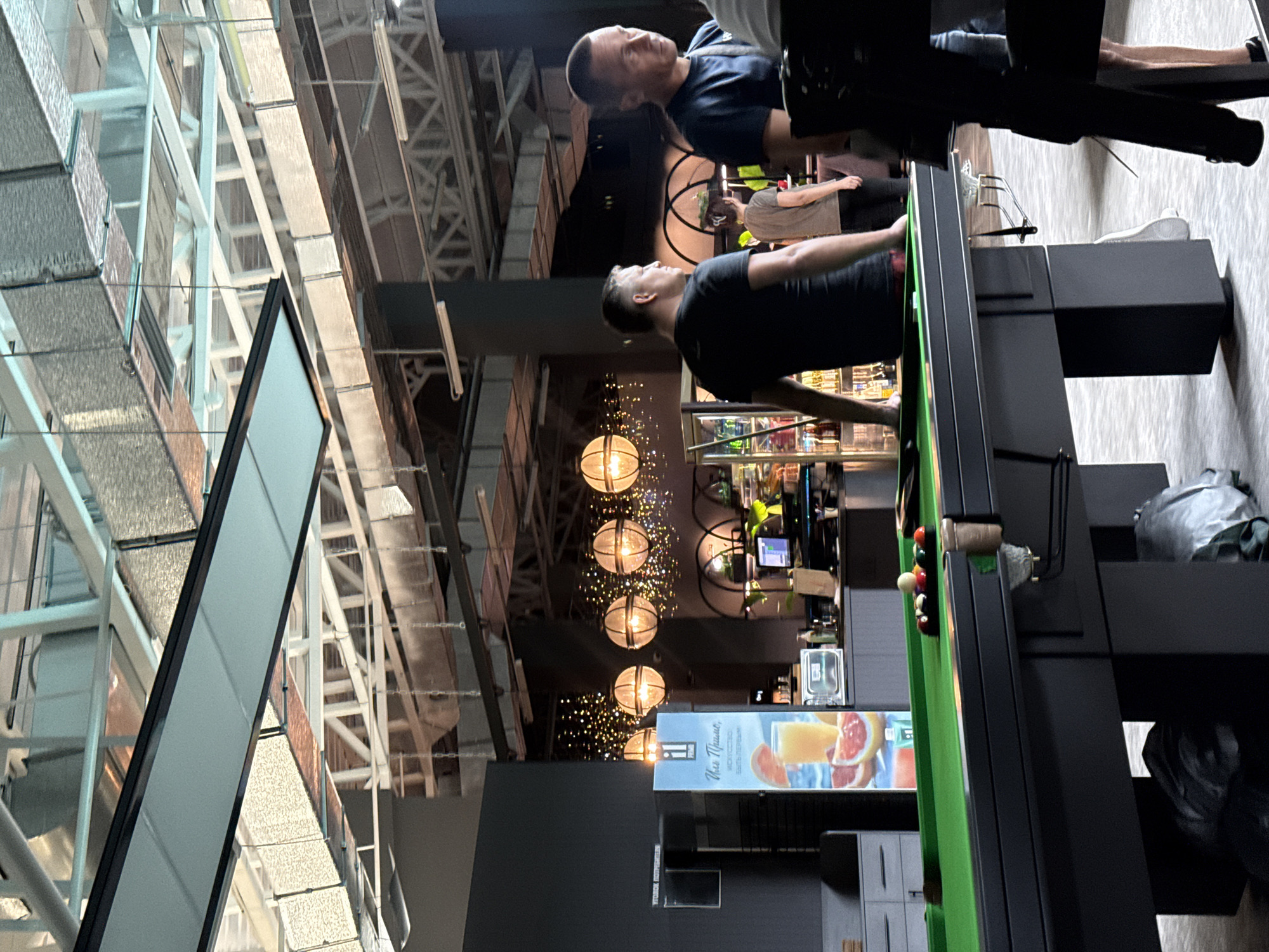The image depicts an indoor scene with a pool table in the foreground. The pool table is green with a set of colored balls arranged on it. To the left of the pool table, there is a display with images of fruit and text in Cyrillic. In the background, several people are visible. One man in a black t-shirt is standing near the pool table, facing away from the camera. Another man in a dark blue t-shirt is standing to the right, looking towards the side. A woman in a grey t-shirt is walking away from the camera in the mid-ground. The environment appears to be a bar or entertainment venue, with decorative string lights and large spherical lamps hanging from the ceiling. The ceiling structure is industrial, with visible beams and ductwork. The overall impression is of a casual social gathering. The provided location is Балаклавский округ, Ukraine, however, this cannot be confirmed or denied from the image alone.