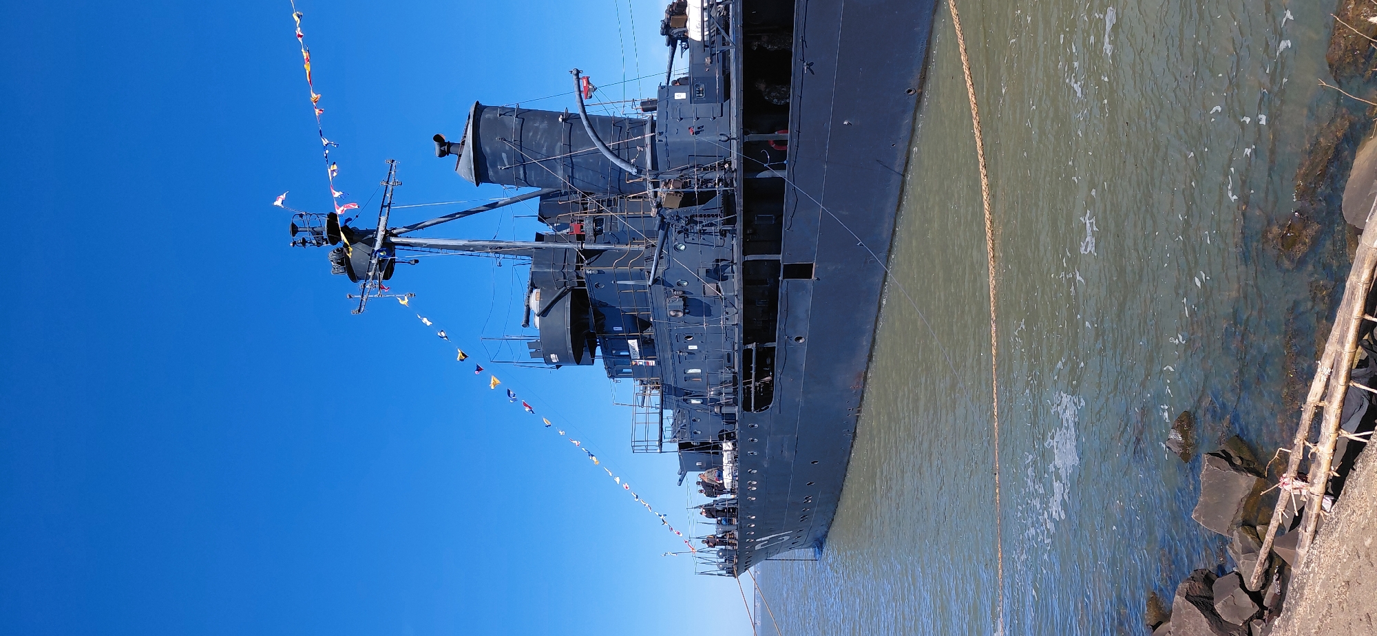 The image shows a large, dark-colored ship docked in murky water, likely the Parana River, near the city of Encarnacion, Paraguay. The ship has a tall mast adorned with a string of international maritime signal flags. Several people are visible on the ship's deck, and some appear to be looking out at the water. The shoreline is rocky, with some debris, including wooden logs, washed ashore. The sky is a clear, bright blue, suggesting a sunny day. The ship is likely a historical vessel, possibly a museum ship, given its stationary position and the presence of flags indicating a special occasion or event. The water appears somewhat polluted, with a greenish-brown hue and some foam on the surface. The overall scene depicts a serene yet somewhat industrial maritime environment.