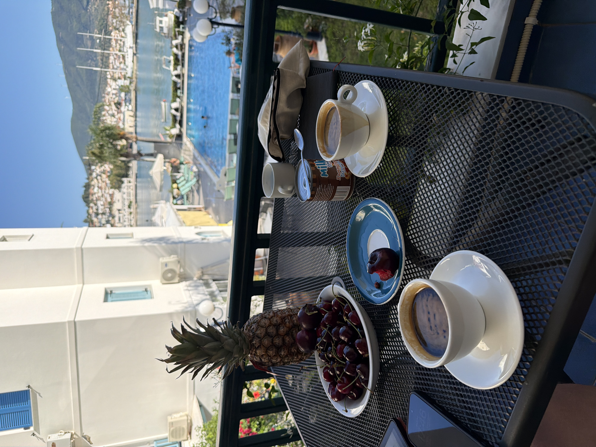 The image presents a vibrant scene from a balcony overlooking a picturesque coastal town, likely in Poros, Greece. The primary focus is a black mesh metal table set with various breakfast or snack items under a clear, bright blue sky, suggesting morning or early afternoon.
On the table, from left to right, are several items: a white cup with a saucer containing a dark beverage, possibly coffee, and parts of mobile devices. Next to them are a white bowl brimming with dark red cherries and a whole pineapple. A small blue saucer holds a dark plum or cherry that has been bitten, revealing its red flesh and a pit. A brown cylindrical can, partially visible, clearly displays the word "MILK" on its label, indicating it's likely condensed milk. Further to the right, another white cup with a saucer also holds a dark beverage, and behind it, a lighter fabric pouch or cap is visible.
Beyond the dark metal balcony railing, a bright blue swimming pool is visible in the mid-ground, with a few blurred figures enjoying a swim and white loungers arranged around its edges. Below the pool area, several white buildings with distinct blue window shutters and visible air conditioning units dot the landscape, showcasing typical Greek island architecture. In the background, a wide expanse of the sea is dotted with various boats and yachts. A large, verdant mountain range rises prominently on the distant horizon, completing the scenic view. The bright sunlight and clear sky contribute to a pleasant and relaxed atmosphere.