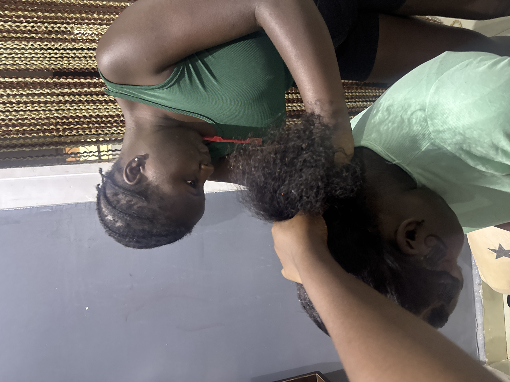In Gura Top, Nigeria, two young Black females are engaged in braiding hair indoors. The individual in the foreground, wearing a light green, ribbed t-shirt, is positioned with their head bowed, receiving the hairstyle. Their dark, curly hair is being manipulated by the second female, who is standing and leaning over. This second female wears a dark green, ribbed tank top and dark shorts. Her hands are actively working on the hair of the person in front of her. The background features a grey wall and a beaded curtain in shades of brown and yellow. The lighting appears to be artificial, creating a slightly dim atmosphere.