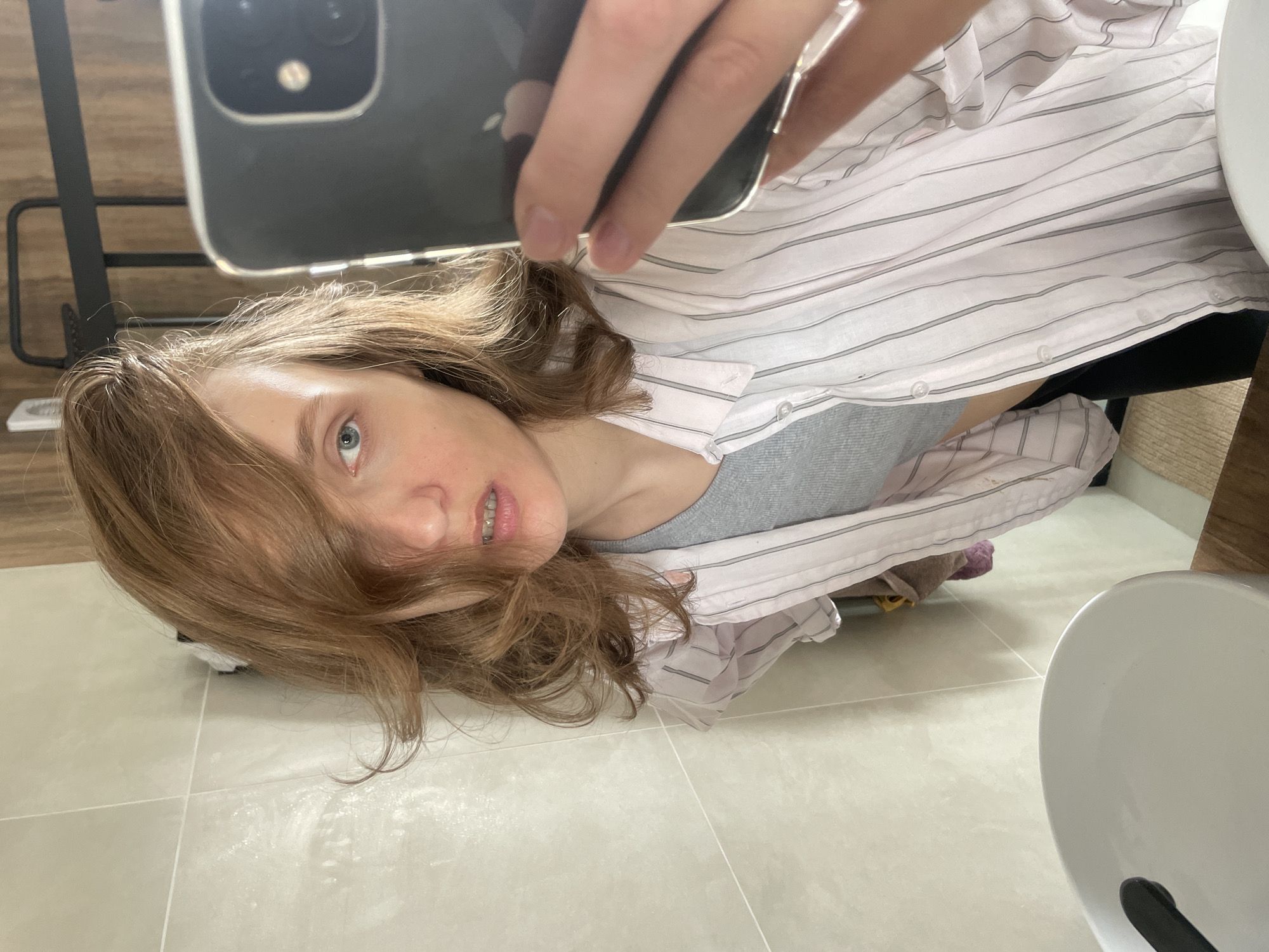 A person is taking a selfie in what appears to be a bathroom. The individual has light brown hair, blue eyes, and braces. They are wearing a striped button-down shirt over a gray tank top. The background includes a light-colored tile floor, a white sink or toilet bowl, and a wooden cabinet. The exact location in Подшивалово, Russia cannot be definitively determined from the image.