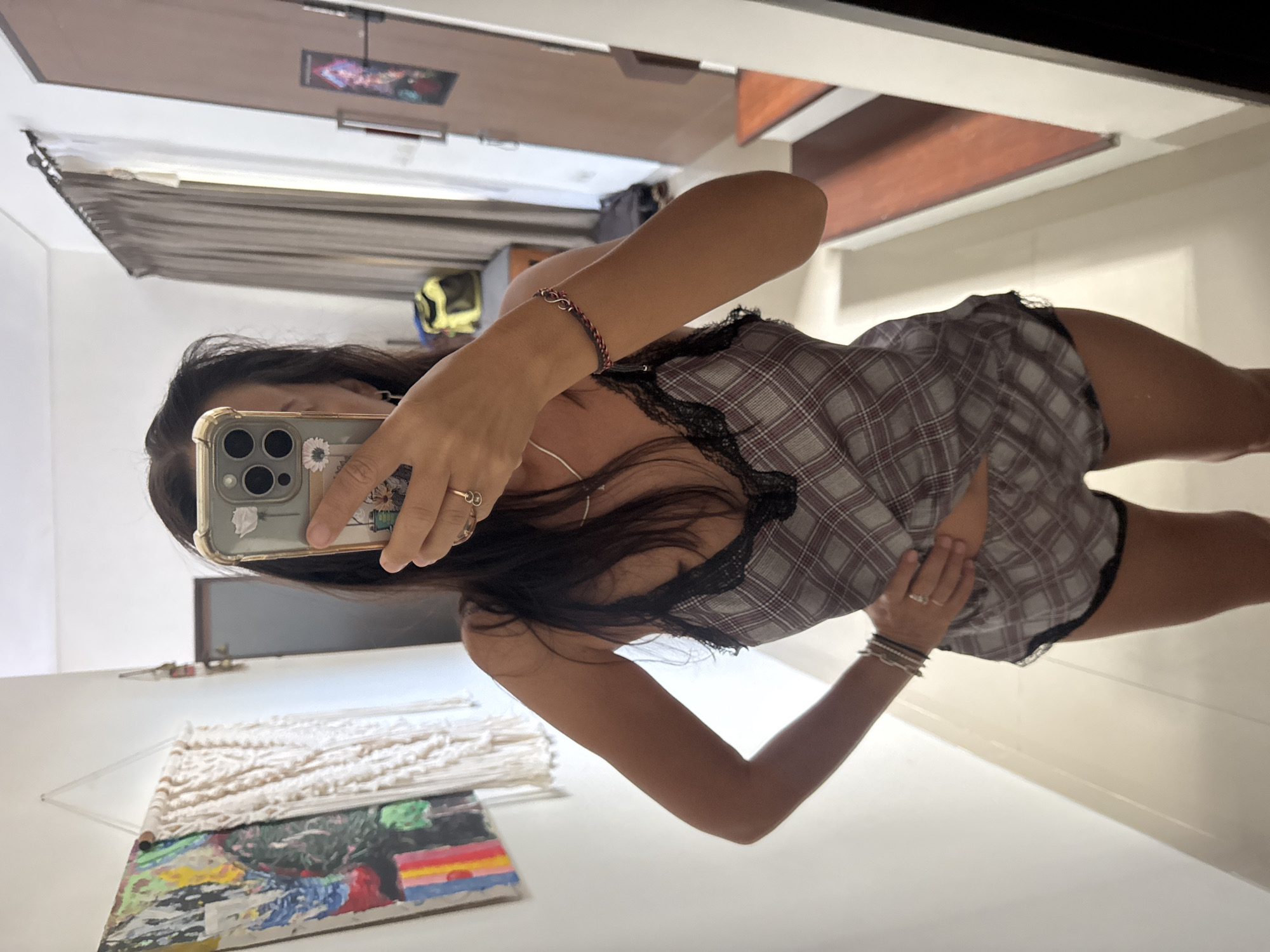The image shows a woman taking a selfie in a room. She is wearing a plaid pajama set with lace trim. In the background, there is a door and a window. A woven wall hanging with paint on the bottom is visible in the foreground. The location is inferred to be in Kuta Selatan, Indonesia.