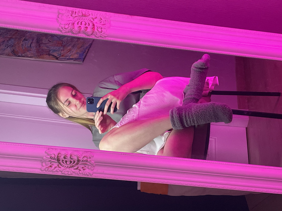 A woman is reflected in a pink-lit mirror, holding a blue phone and looking at the screen. She wears a grey shirt and a white nightgown, with grey fuzzy socks on her feet. A painting hangs above her, partially visible in the mirror. The mirror's ornate frame is also bathed in the pink light.