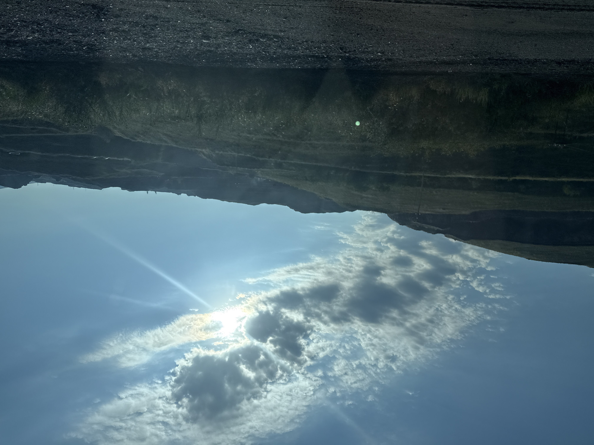 The image depicts a scenic view of a sunlit sky with scattered clouds over a hilly terrain. The sun's rays pierce through the clouds, creating a bright, lens flare effect. The landscape appears to be a quarry or mine, with terraced slopes and sparse vegetation. The foreground is a dirt road or path, covered with gravel and small stones. The sky is a vibrant blue, with the sun casting a warm glow on the scene. The location is identified as the city of Arzni, Armenia. There are no people or specific objects of interest visible in the image, other than the natural elements of the landscape and sky.