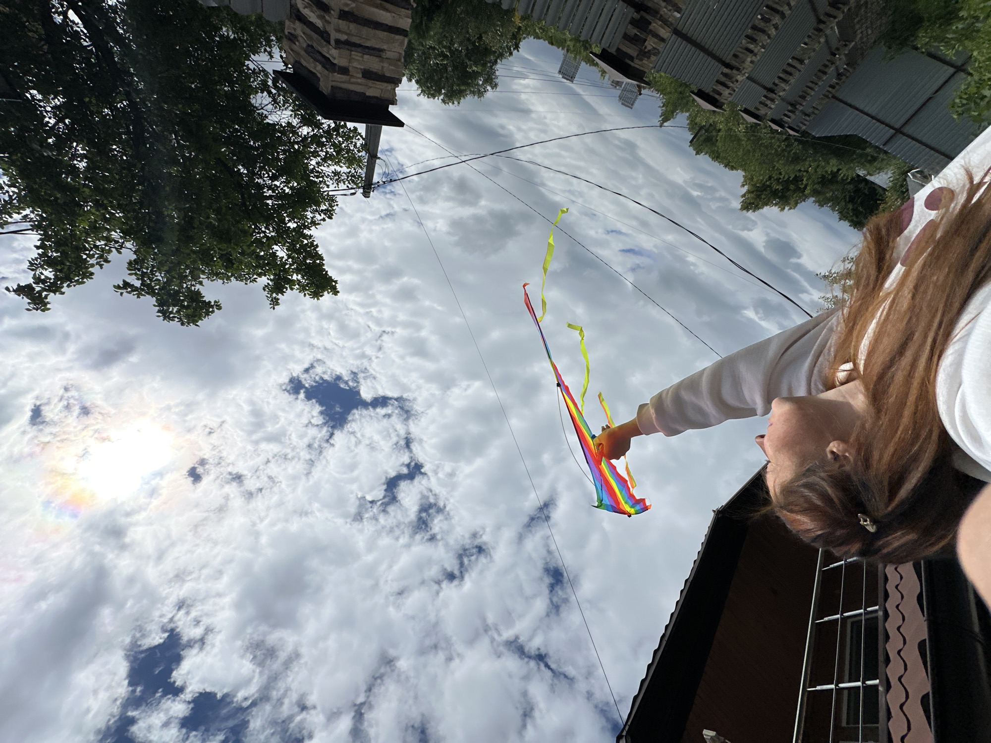 A young woman with long, brown hair is holding a rainbow-colored kite with yellow streamers against a cloudy sky. She is looking upwards, with the kite held high in her outstretched hand. The kite appears to be in mid-air, with the streamers fluttering. The background shows a suburban or rural setting, with a house and trees visible. The overall impression is one of freedom and joy, with the bright colors of the kite contrasting with the overcast sky. The image seems to be taken outdoors on a day with a mix of sun and clouds. The orientation of the image is somewhat unusual, as if taken from a low angle looking up. No specific details about Nizhny Novgorod, Russia, are visible in the image.