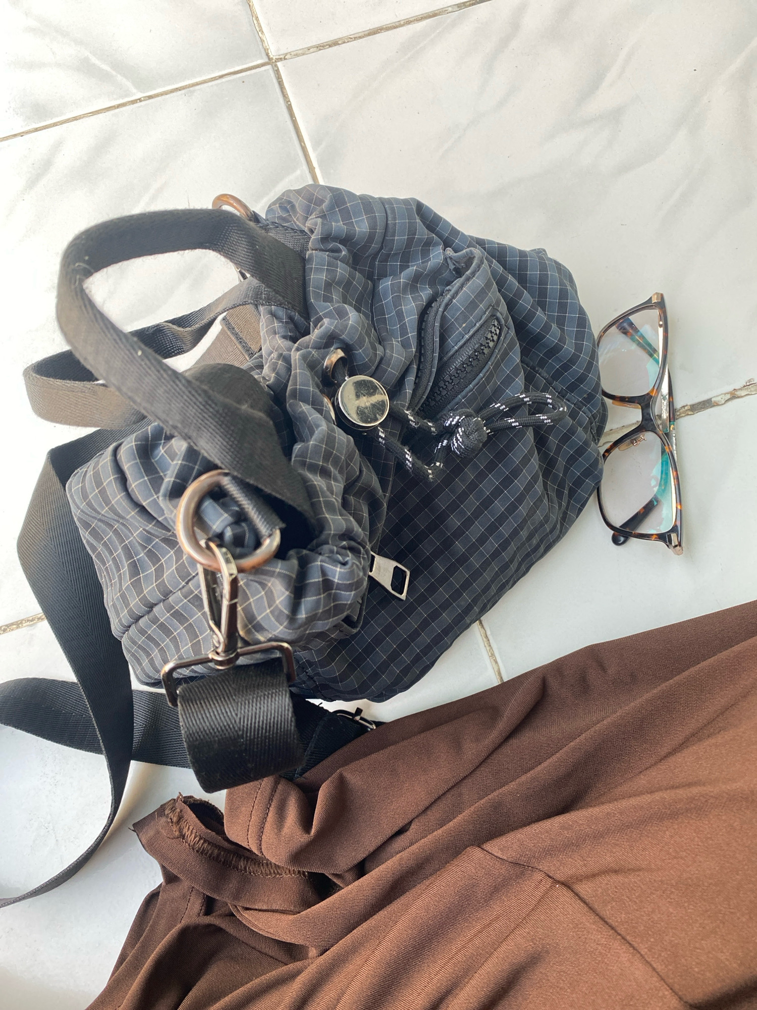 An overhead shot shows a black and grey checkered drawstring bag with a black strap and gold hardware, placed on a white tiled floor. Next to the bag are a pair of tortoiseshell glasses. To the right of the bag is a crumpled brown garment. The scene is indoors, possibly a home, in the city of Kedung Sukodani, Indonesia.