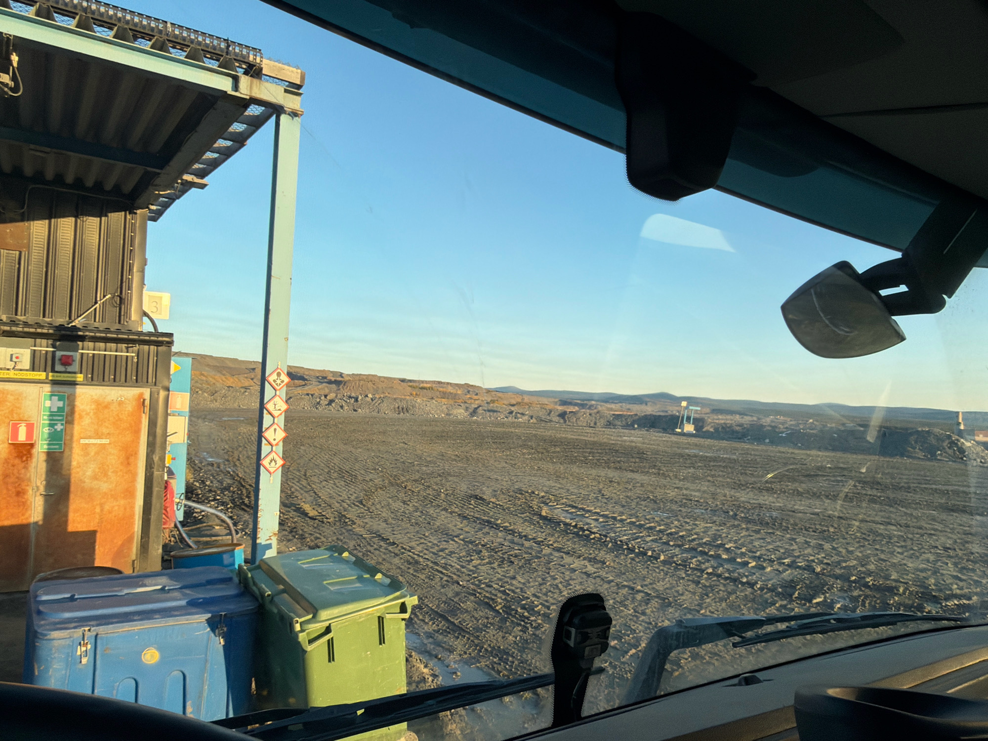 The image captures a view from inside a vehicle, looking out at an outdoor industrial site under a clear blue sky. In the foreground, the dashboard of the vehicle is visible, along with a windshield wiper and a rearview mirror. To the left, a structure with a corrugated metal roof and a light blue support pillar is present. Adjacent to this structure are two large bins, one blue and one green. Several warning signs are attached to the blue pillar, including hazard symbols.
The midground consists of a vast, open area of dark, uneven terrain with visible tire tracks, suggesting recent vehicle activity. In the distance, a hilly landscape with sparse vegetation and some man-made structures can be observed. The overall impression is one of an active industrial or mining operation in a somewhat barren, possibly cold, environment. Based on the presence of Norwegian text on the warning signs, such as "NØDSTOPP" (Emergency Stop), and the industrial context, the location is likely within the industrial area of Gällivare, Sweden, which has significant mining operations.