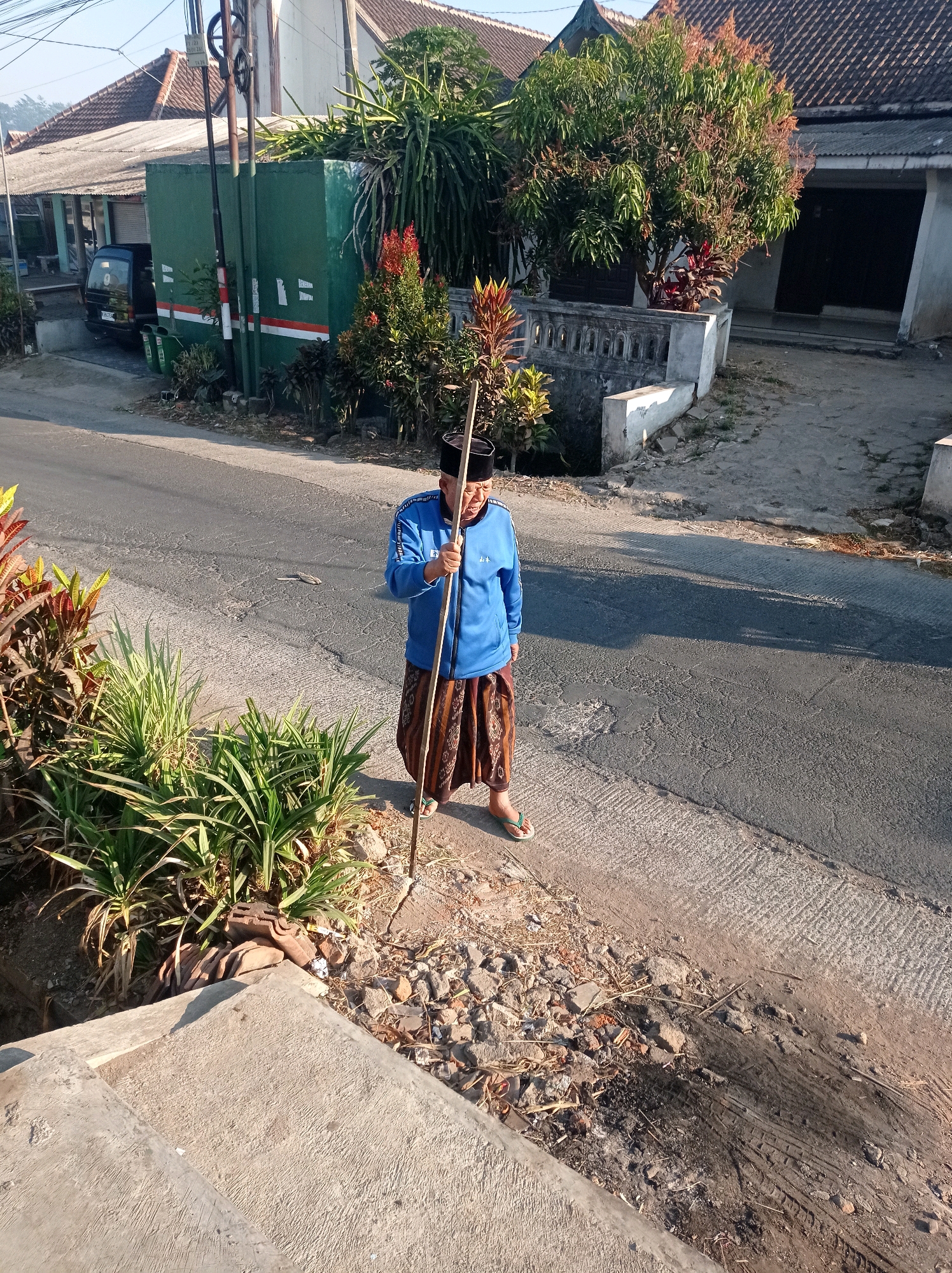 A man stands on the side of a paved road in Purwosekar, Indonesia, holding a long wooden pole. He is wearing a blue long-sleeved shirt with white trim and a patterned sarong. On his head, he wears a black cap. The man is looking towards the camera. Beside him, there is a patch of green plants and scattered debris. The road is made of asphalt and shows signs of wear and tear. In the background, there are houses with tiled roofs, some vegetation, and a green utility box with white markings. A black vehicle is parked in front of one of the houses. The sun is shining, casting shadows on the road.