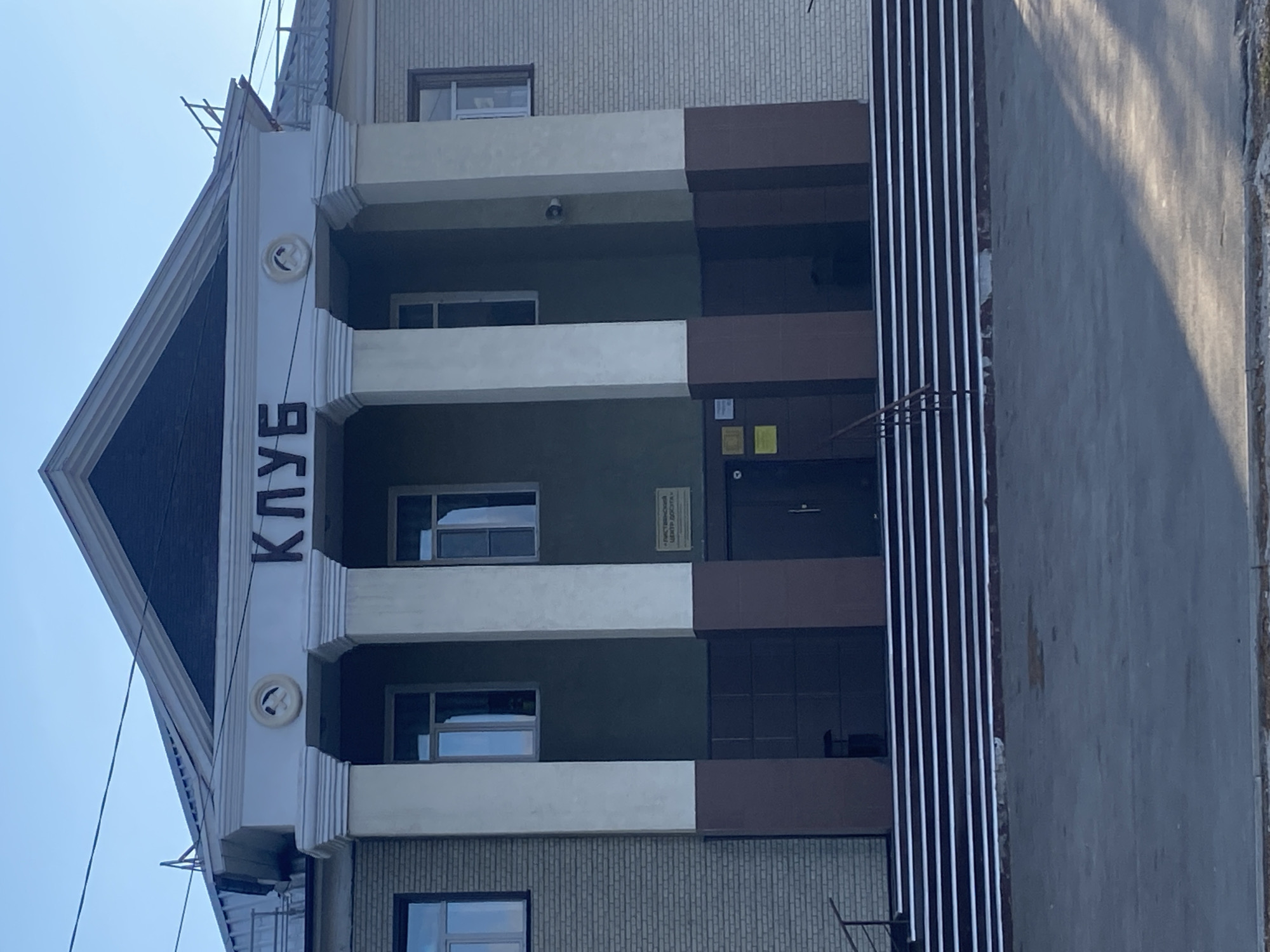 The image shows the facade of the "Club" building in Listvyanskiy, Russia. The building has a classical architectural style with four large white columns supporting a pediment. Above the columns, in large dark letters, is the word "КЛУБ" (Klub), meaning "Club". On either side of the word, there are circular decorative elements, each containing a hammer and sickle emblem. The building has a light brick exterior on the sides, with dark brown and gray paneling on the lower sections of the facade. Several windows are visible on the upper floors. In front of the building, there is a wide set of stairs with white and dark horizontal stripes. A metal railing is present on the left side of the stairs. The ground in front of the stairs is paved. The sky is clear and blue.