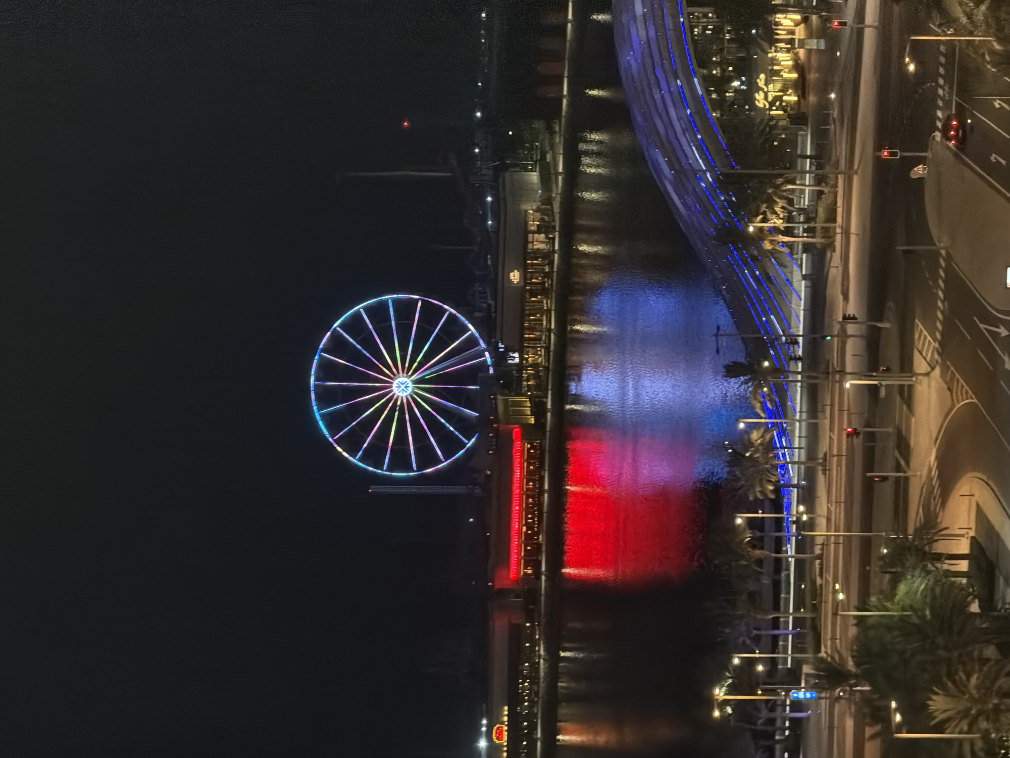 An aerial night-time shot captures a brightly lit Ferris wheel, its spokes adorned with a spectrum of vibrant colors, including pink, purple, blue, green, and yellow, against a dark sky. Below, a body of water reflects the illuminated structures along its bank. A building facade glows red, casting a vivid reflection on the water's surface, with streaks of blue light also visible. Palm trees are scattered along the waterfront, and a modern, arched structure with blue lights runs across the foreground. A street scene with traffic lights and roads is visible at the bottom of the frame, with a red car present.