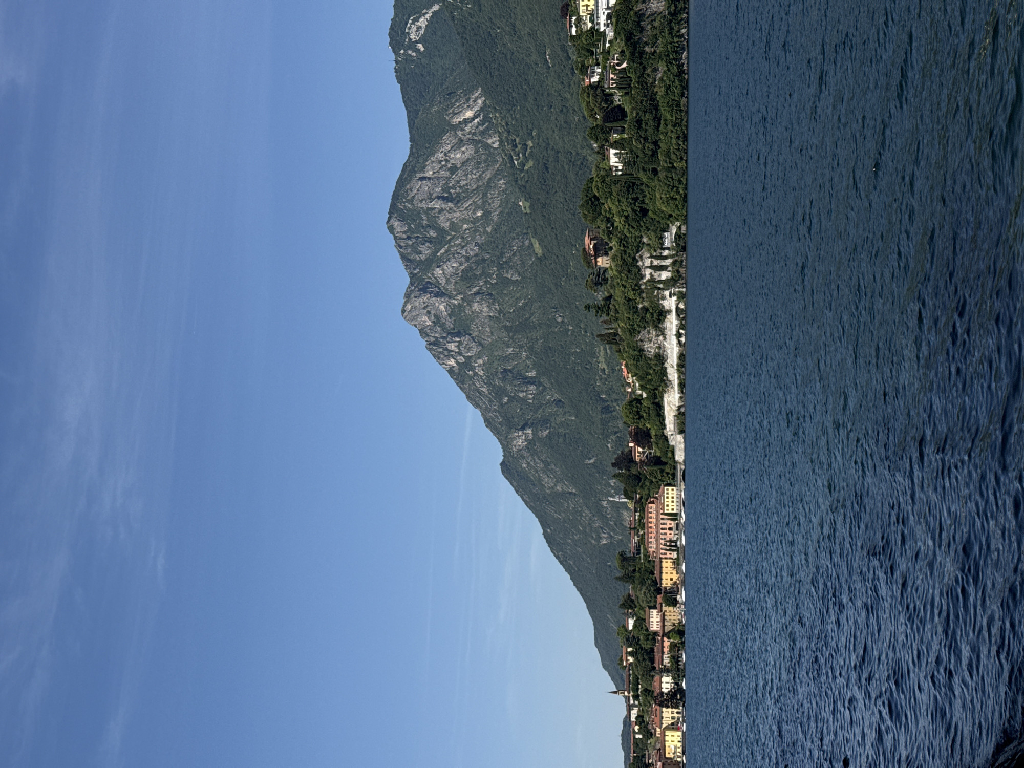 This is a landscape photograph taken from a body of water, likely a lake, looking towards a mountainous, wooded shoreline dotted with buildings. The image is oriented vertically, with the sky occupying the left half and the mountain and water occupying the right half.
The sky is a clear, bright blue with a few wispy white clouds on the left side. The right side of the image is dominated by a steep, rugged mountain face covered in dark green vegetation. Nestled on the lower slopes of the mountain and extending down to the water's edge is a picturesque village. The buildings are varied in color, with many featuring pastel hues of yellow, pink, and orange, and are clustered together along the shoreline. Some appear to be houses, while others could be hotels or other structures. The presence of small boats docked along the shore suggests a popular tourist destination.
The water is a deep blue and appears slightly choppy, with ripples indicating a gentle breeze. The sunlight suggests it is daytime, likely late morning or early afternoon, given the bright illumination and clear sky. The overall mood is serene and picturesque, evoking a sense of natural beauty and a charming European village setting. No visible text is present in the image.