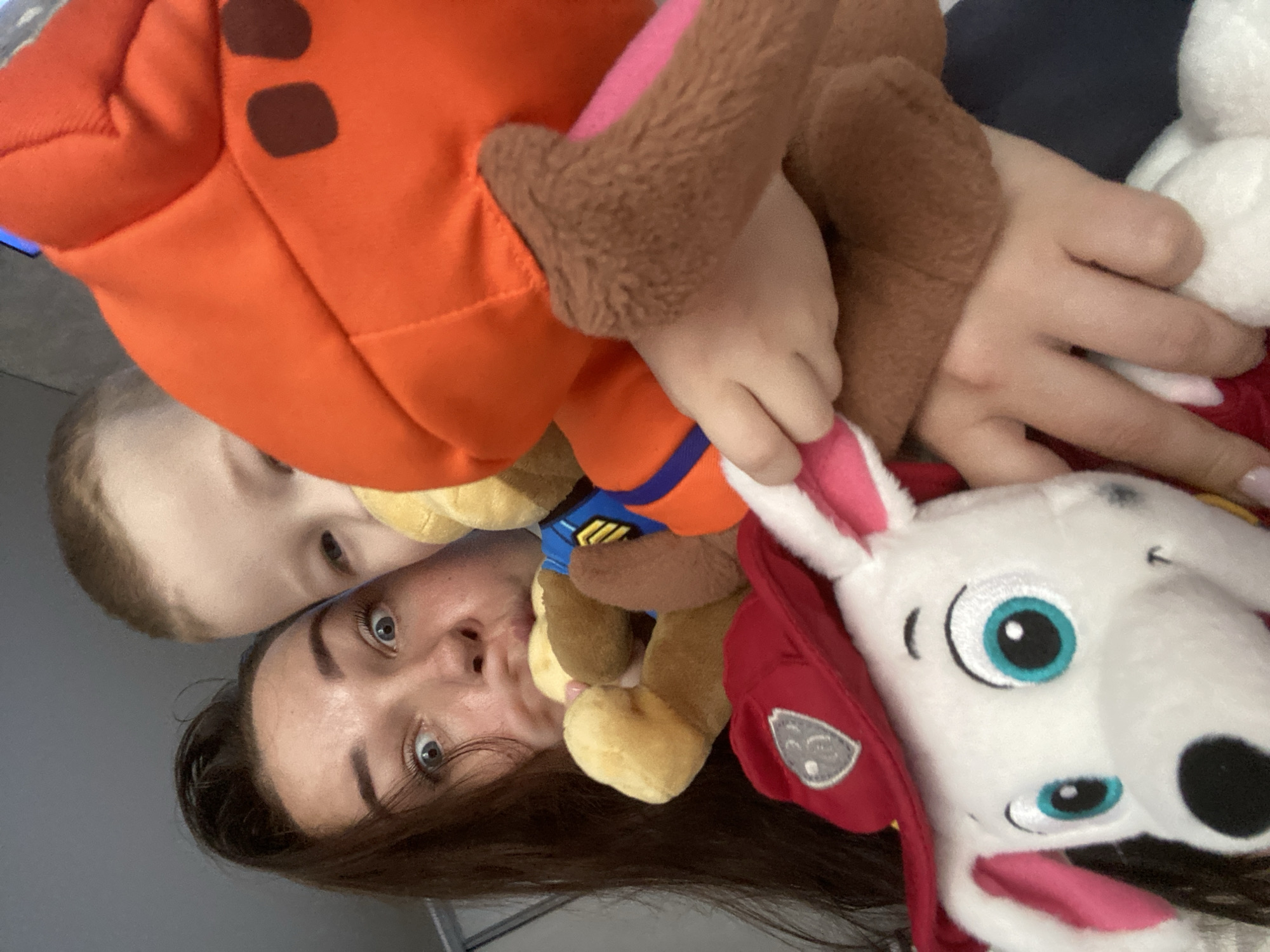 A close-up, canted-angle image shows a woman and a child posing with three Paw Patrol plush toys. The woman is in the foreground, with her head oriented toward the lower part of the frame. Her dark brown hair is visible at the bottom, and her face is angled upwards, revealing her eyes and nose. The child is positioned slightly above her head, also looking towards the camera. The three plush toys are arranged around them, with an orange and brown toy at the top, a yellow toy near the center, and a white and red toy on the right side of the frame. There is no clear indication of the location being Sochi, Russia. The background is a neutral gray color.