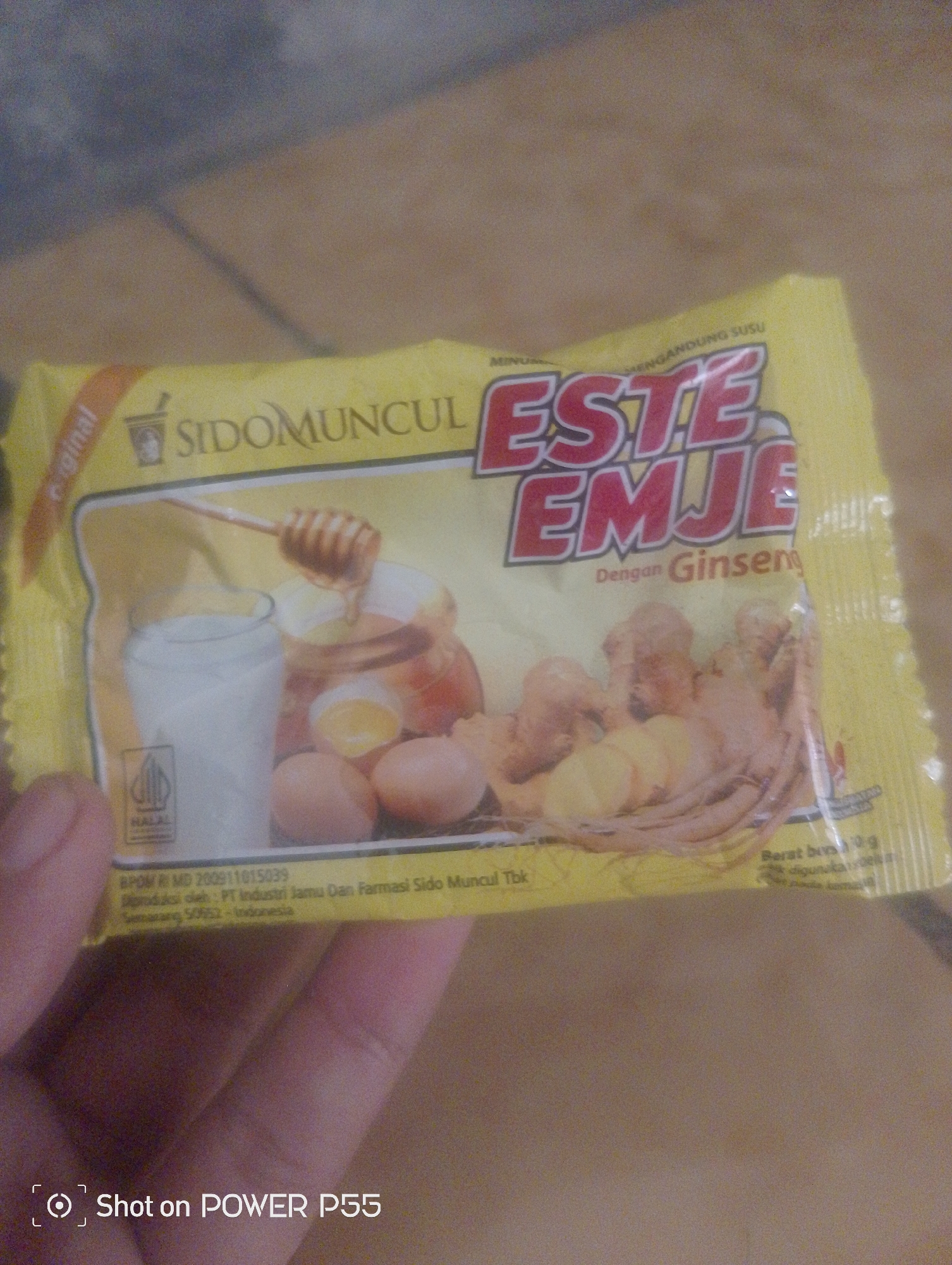 A yellow sachet of Sido Muncul Este Emje with Ginseng is held by a person's hand. The sachet features an illustration of a glass of milk, a honey dipper with honey, cracked eggs, and ginseng roots. Text on the sachet indicates it contains milk and ginseng. The background is out of focus and appears to be a tiled or concrete surface. There is no visible information to suggest the location is in Lebaksiu, Indonesia.