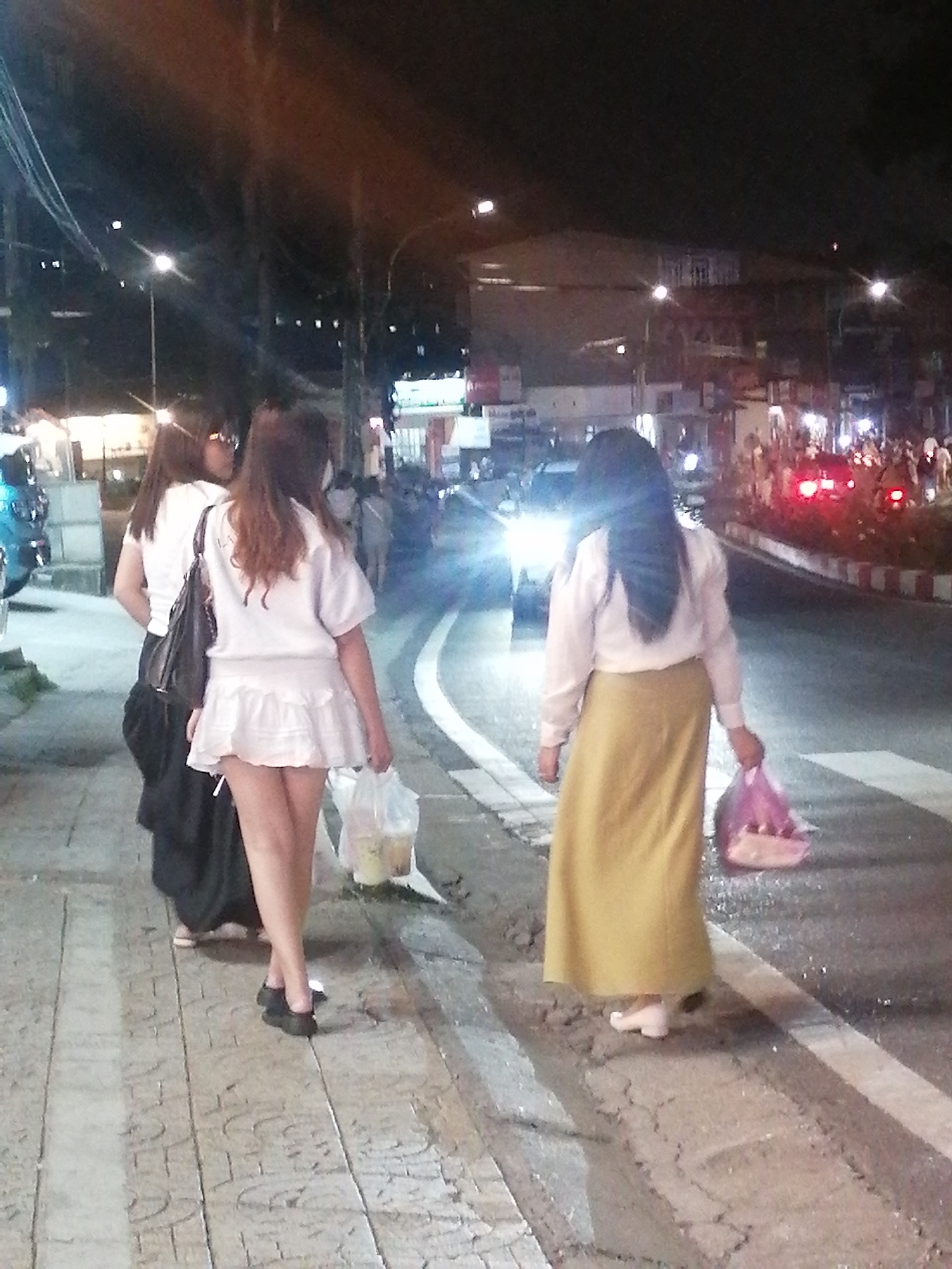 The image captures a nighttime scene on a city street, potentially in Da Lat, Vietnam, characterized by streetlights and illuminated buildings in the background. In the foreground, three individuals are walking along a paved sidewalk. The person closest to the viewer is a woman wearing a long, mustard-yellow skirt and a light-colored long-sleeved shirt, carrying a pink plastic bag in her right hand. To her left, another woman is walking in the same direction, dressed in a short white ruffled skirt and a white t-shirt, with a dark bag slung over her shoulder. Her legs are crossed as she walks. A third woman, partially obscured, is walking ahead of them, wearing a dark skirt and a white top, with a dark bag on her shoulder. In the background, vehicles with their headlights on are visible on the road, along with distant lights and structures suggesting urban activity. The sidewalk appears to be made of textured paving stones, and the road has a white line marking its edge.