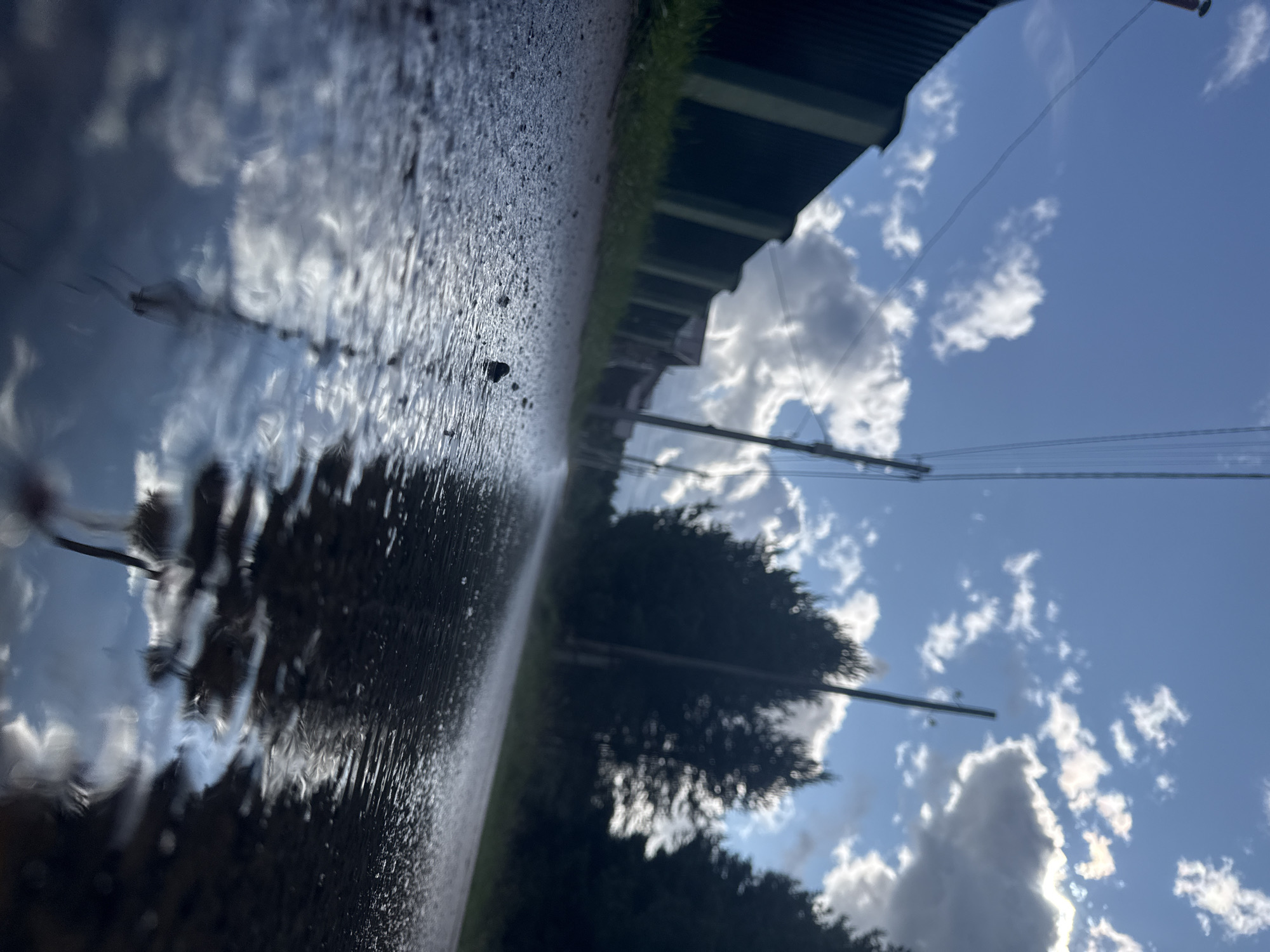 The image depicts a low-angle, wide shot of a wet, gravelly road after rain. The foreground is dominated by the glistening road surface, reflecting the sky and surrounding elements. Several small pebbles are visible on the road. In the middle ground, a dark green fence runs along the left side of the road, bordering a grassy area. Beyond the fence, houses and other structures are partially visible, blurred by the focus. Several utility poles with overhead wires stretch diagonally across the frame. On the right side of the road, silhouetted trees rise against a bright, partly cloudy sky. The sky is a vibrant blue with scattered white and grey cumulus clouds, some of which are backlit by the sun, creating bright highlights. The overall impression is one of a rural or semi-rural environment in Chaikovsky, Russia, likely on a street or pathway with a rough surface. No people are visible in the scene.