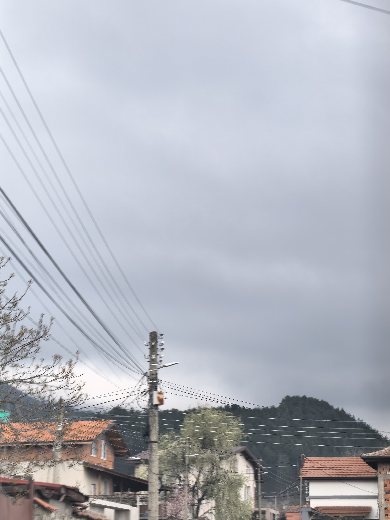 The image depicts a street scene in an urban area with residential buildings and utility poles under an overcast sky. The houses have orange-tiled roofs and light-colored facades. Power lines are strung across the view, obscuring parts of the landscape. A hill covered with trees is visible in the background. The general location is Unknown, Unknown.