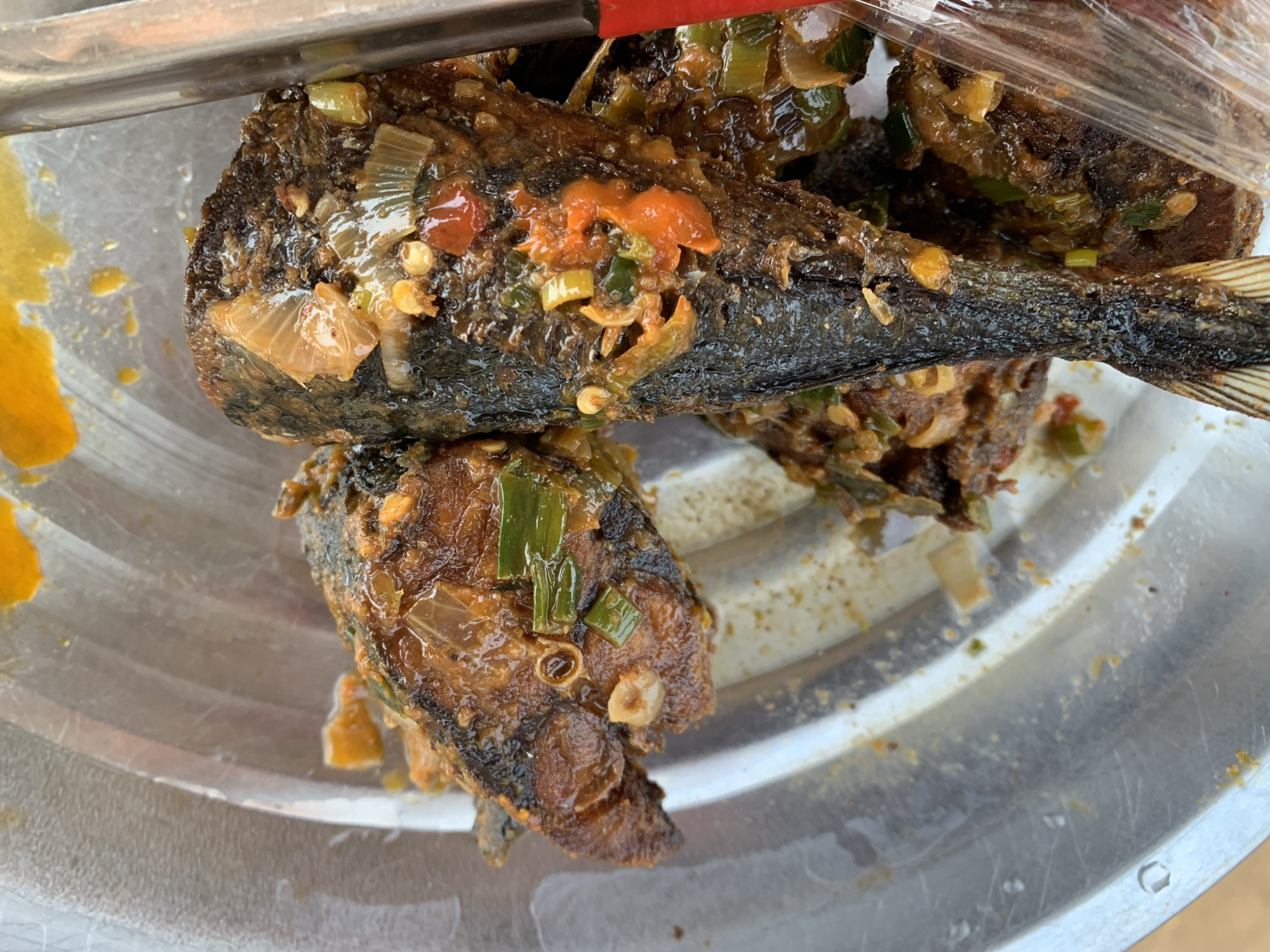 The image shows a plate of fried fish, likely catfish, covered in a dark sauce with visible pieces of green onions, red peppers, and possibly garlic. The fish is whole, with the head and tail visible. The plate is made of aluminum, with some yellow oil or sauce pooled on the side. The context suggests the location could be Rafin Par, Nigeria, although there is no clear visual confirmation of the place.