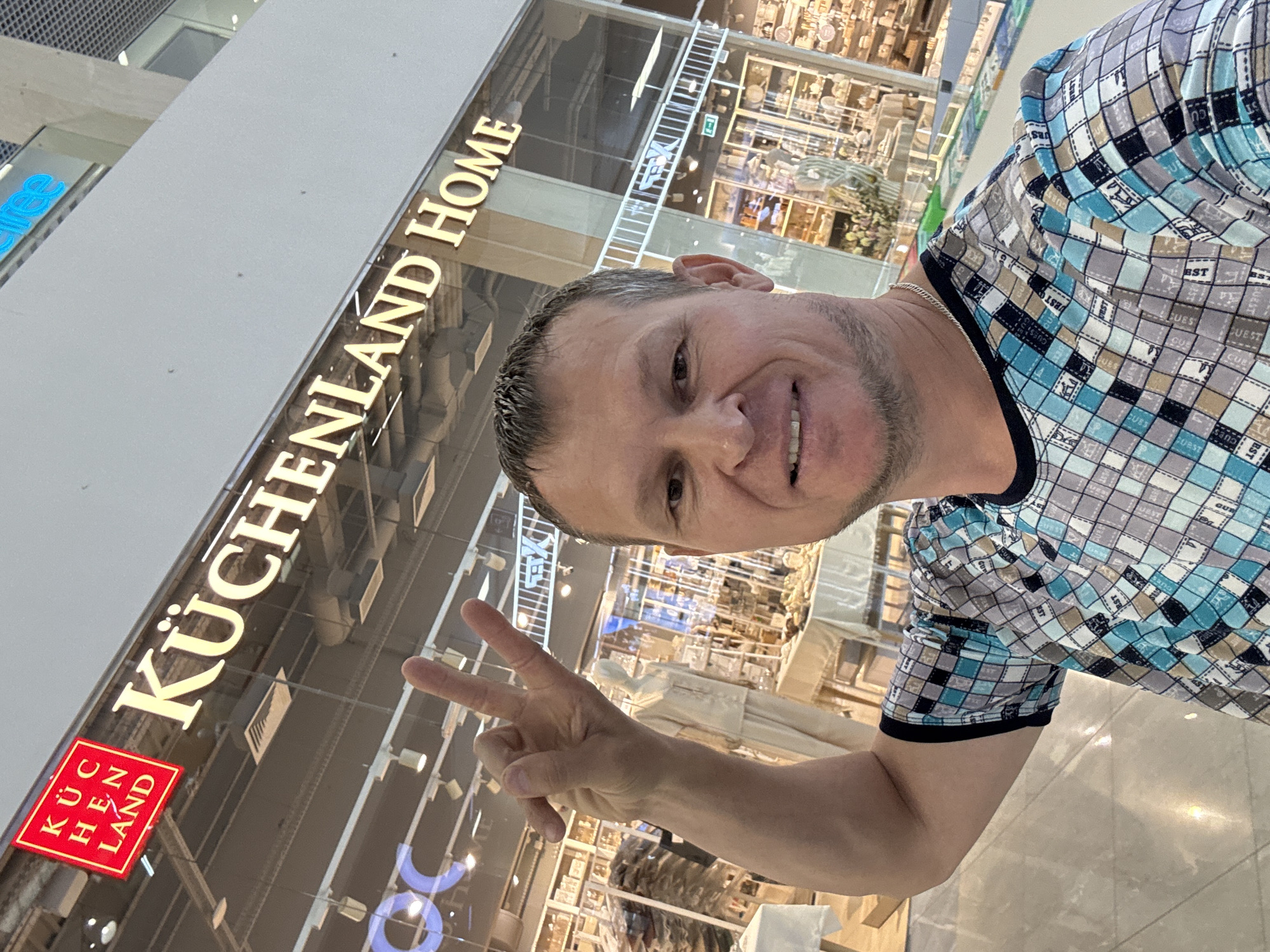 A man, appearing to be of Caucasian ethnicity and in his late 30s or early 40s, takes a selfie in front of a store. He has short, dark hair, a light stubble, and a noticeable scar above his right eye. He is wearing a checkered t-shirt with blue, gray, and tan squares. His left arm is raised, with his index and middle fingers extended in a peace sign.
Behind him, the store's signage reads "KÜCHENLAND HOME" in large, illuminated letters. To the left of this main sign, a smaller red square sign displays "KUC HEN LAND" in white letters. The store appears to be a retail establishment, with displays of goods visible through the large windows. In the background, the interior of the store shows shelves stocked with items, a mannequin dressed in light-colored clothing, and tables with merchandise. The environment suggests an indoor shopping mall, with a brightly lit ceiling and a reflective floor. The overall setting is consistent with the interior of the "Kuchenland" store located in the "Emerald City" mall in Oskemen, Kazakhstan.