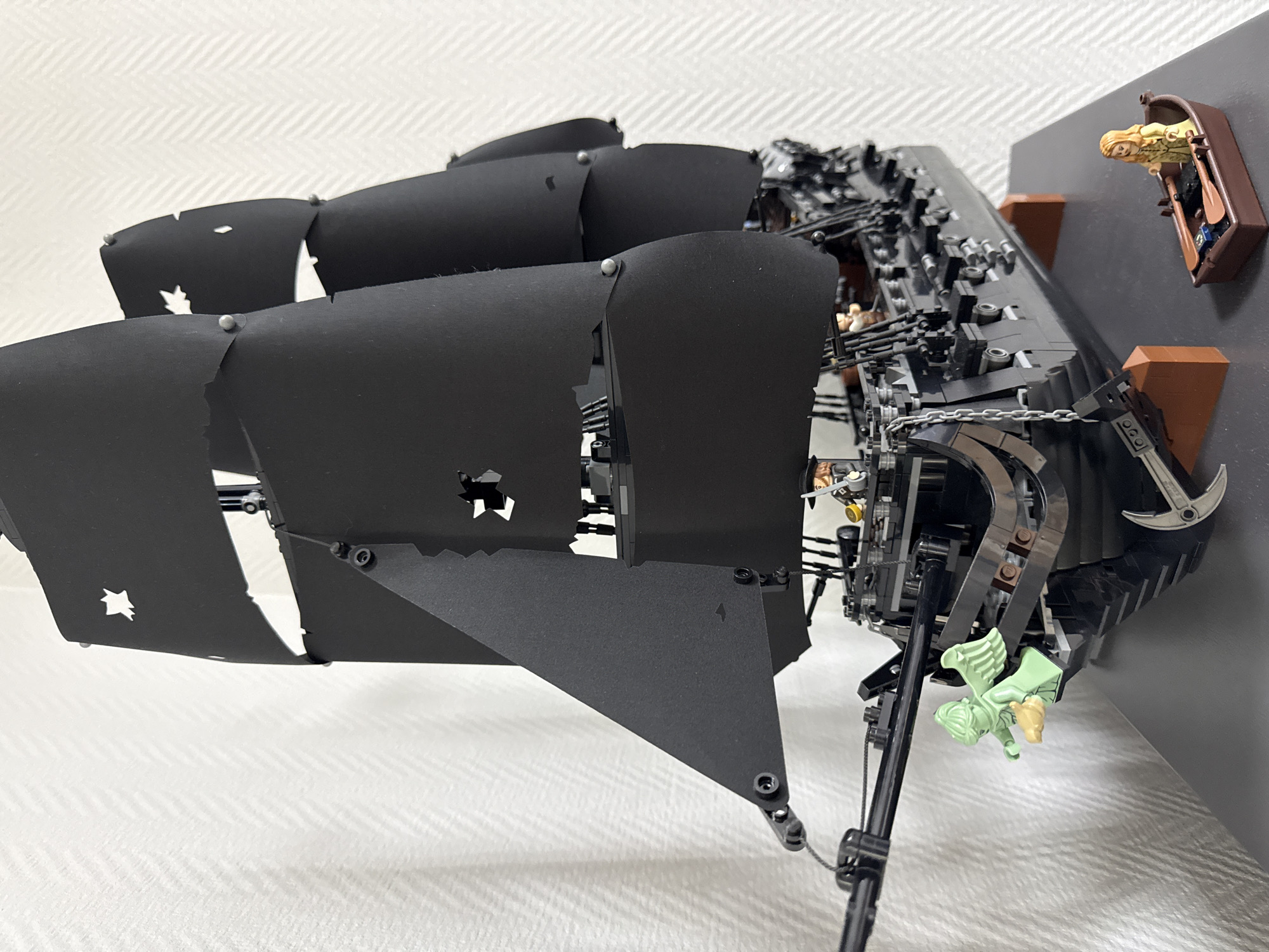 A close-up shot of a partially built Lego ship, primarily black in color. The ship has several black sails with distinctive cut-out shapes. The hull is composed of many small black Lego pieces, and there are brown elements as well. There is a figurine of a woman in a boat with oars. There is a green angel figure on the front of the ship. The model sits on a white surface with a wall in the background. There is no evidence of Krasnoyarsk, Russia.