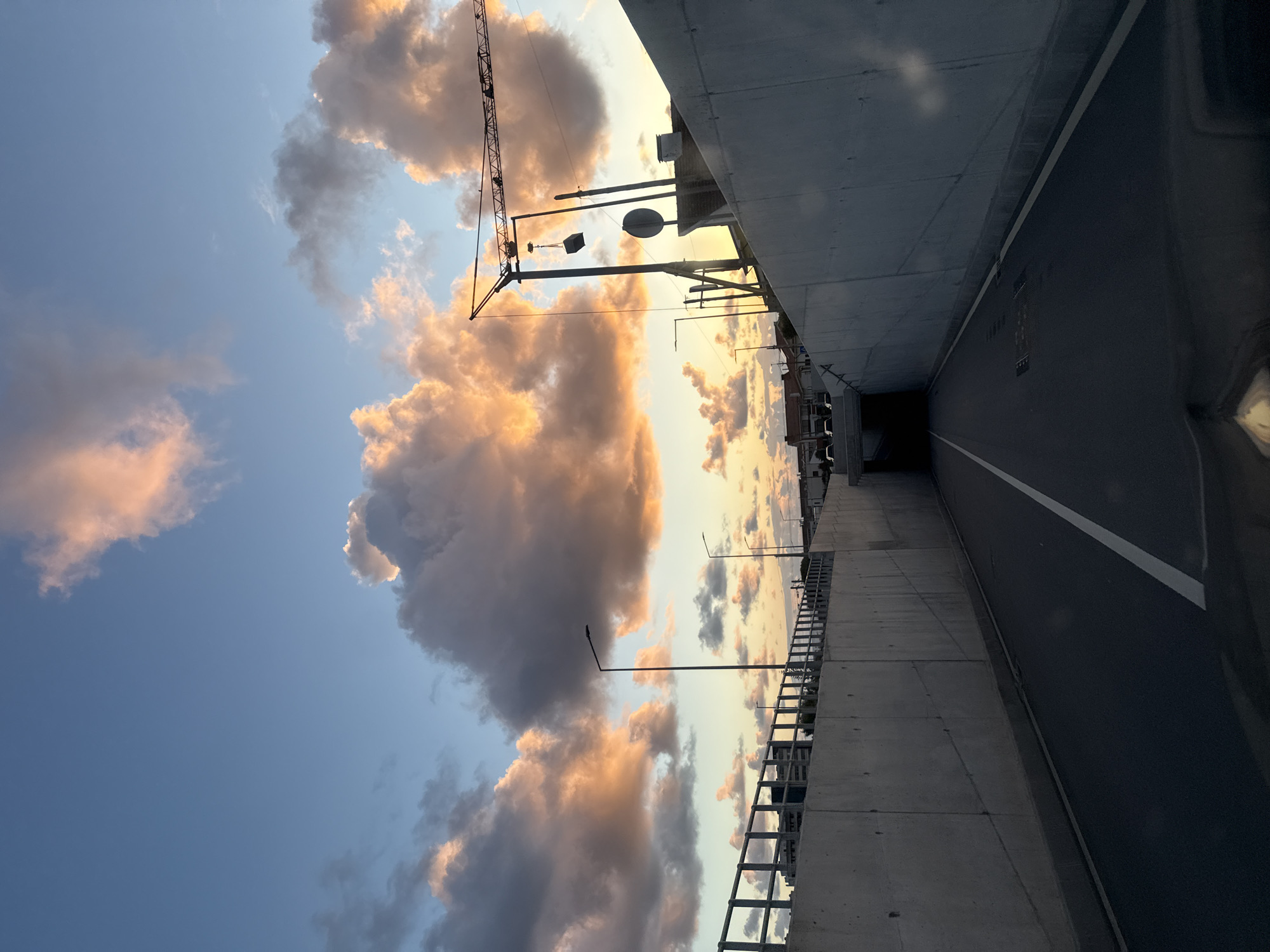 The image depicts a scene in Porto, Portugal. It is taken from under an overpass, looking out at a sky filled with clouds, some illuminated with the warm colors of a sunset or sunrise. A construction crane is silhouetted against the sky. Below the bridge, there is a highway with a white lane divider. Street lights are interspersed along the railing on the left side of the road, which appears to be a pedestrian walkway. The angle suggests that the photo was likely taken at dusk or dawn due to the sky coloring. The environment is urban, evident by the concrete overpass structure and the presence of a highway. There are no visible people.